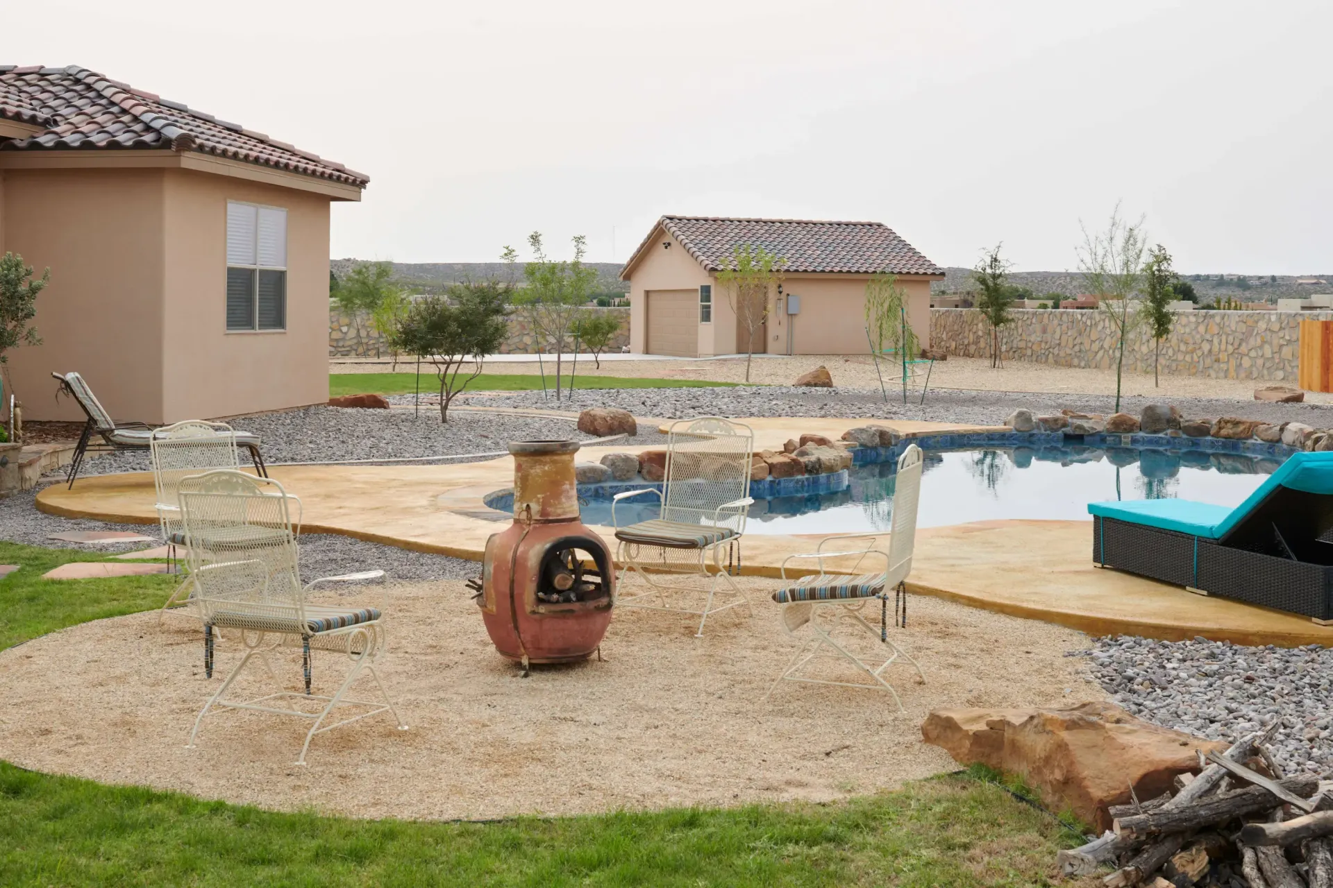 Backyard with a pool, seating, a fire pit, and buildings under an overcast sky.