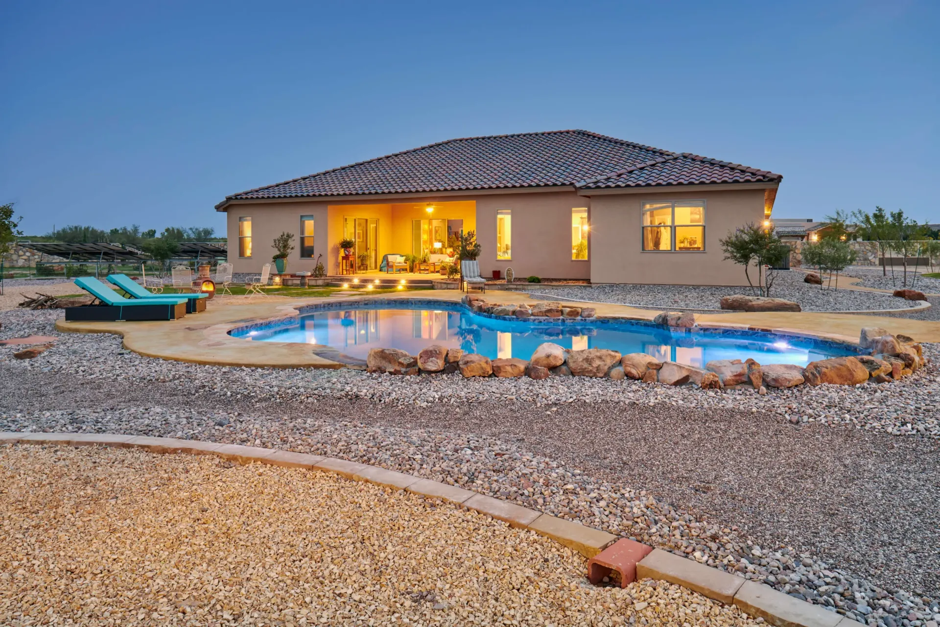 Backyard with pool and house at dusk. Lights illuminate the pool and interior. Ground covered in gravel.