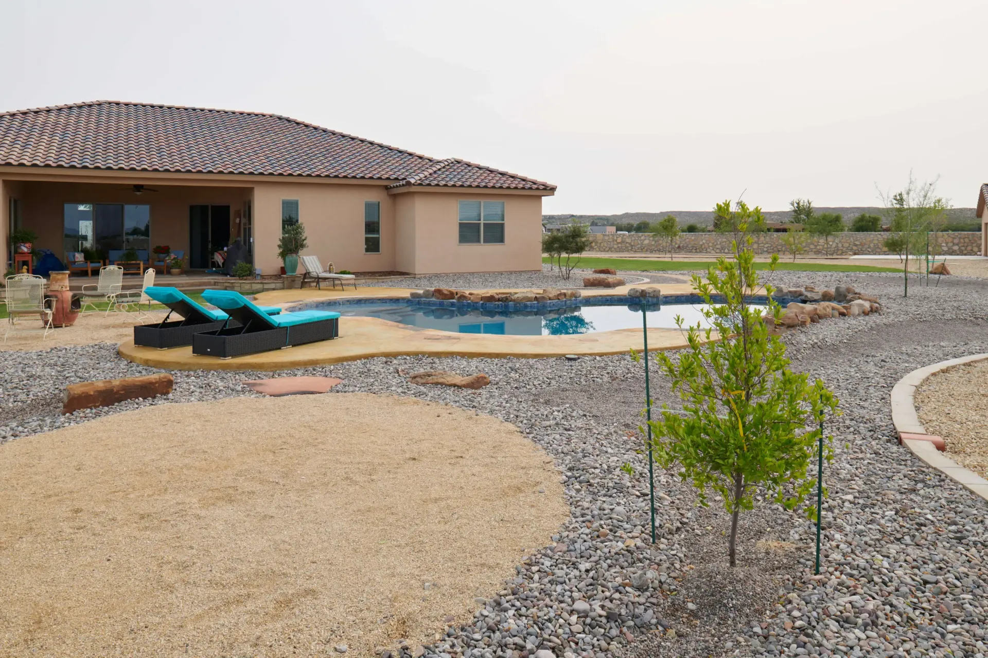 Backyard with a pool and lounge chairs next to a tan stucco house and gravel landscaping.