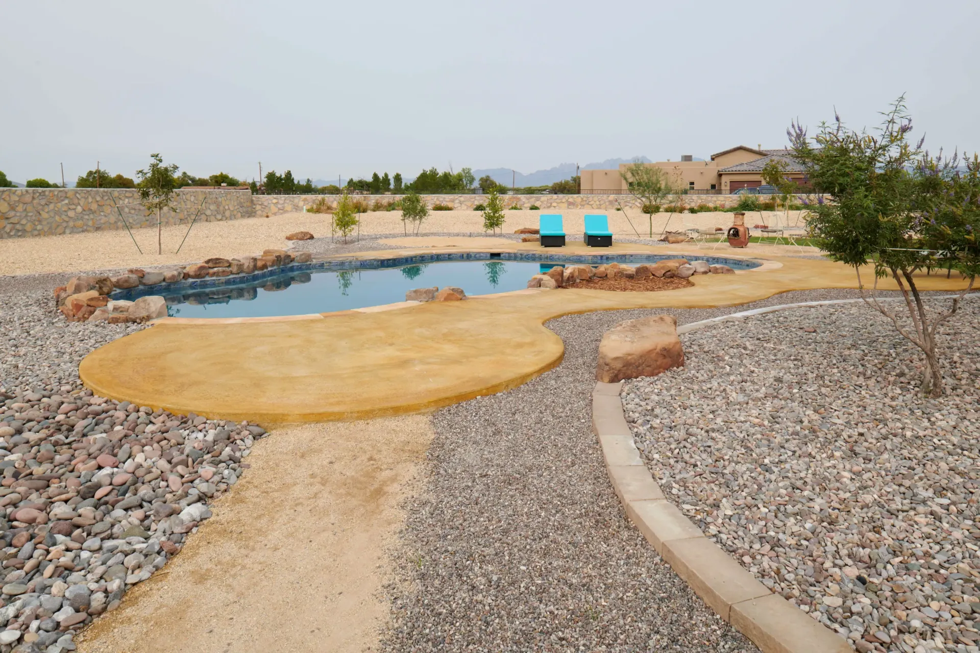A backyard with a natural-looking pool, gravel paths, tan ground cover, and blue chairs.