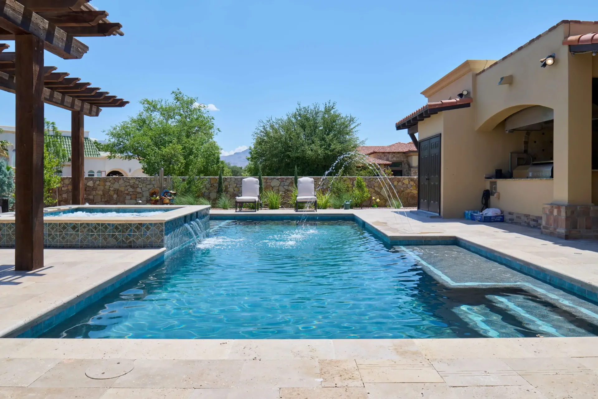 Pool and spa in a sunny backyard with a pergola, landscaping, and a house. Blue water, clear sky.