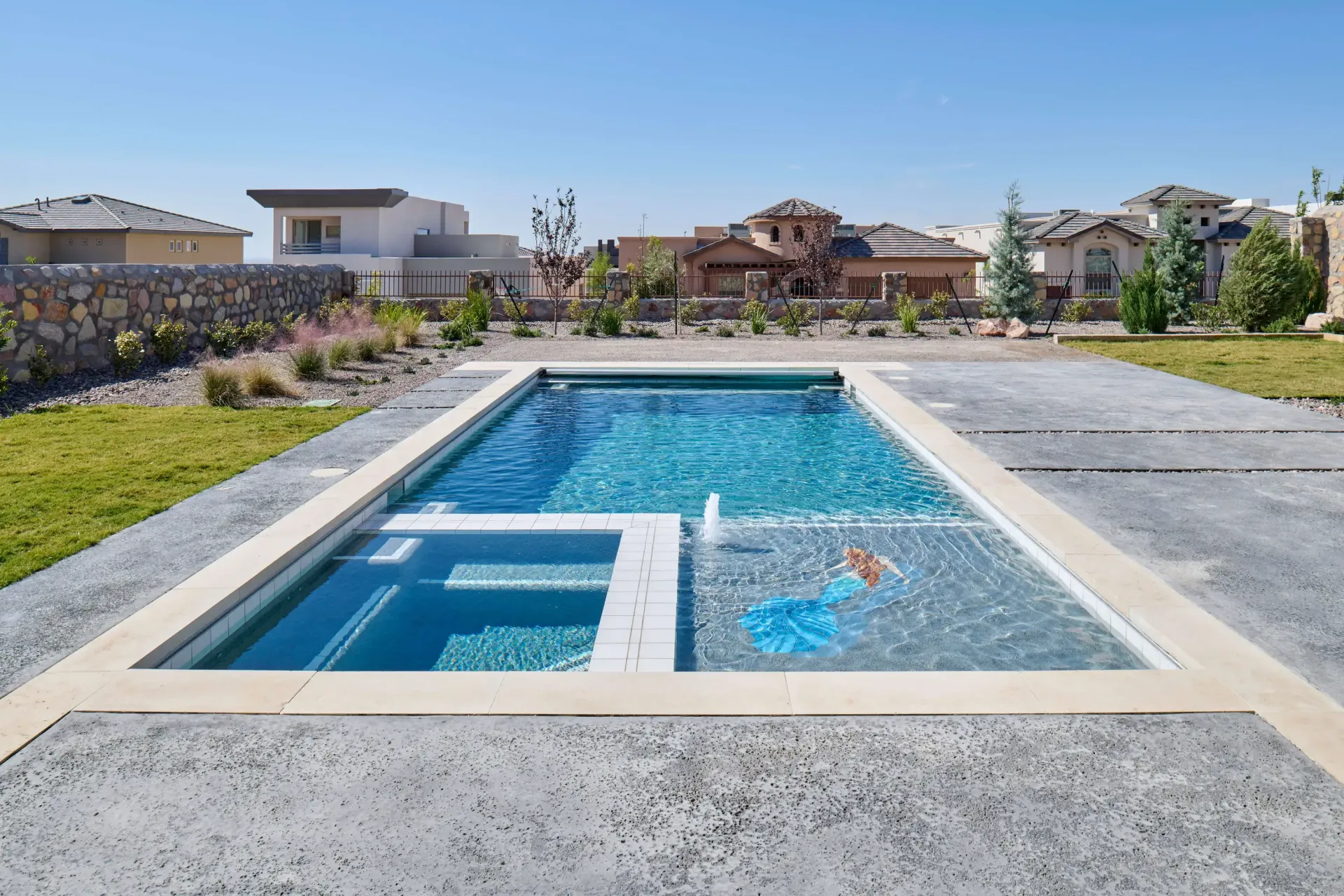 Rectangular pool with attached spa, set in concrete, houses in background under blue sky.