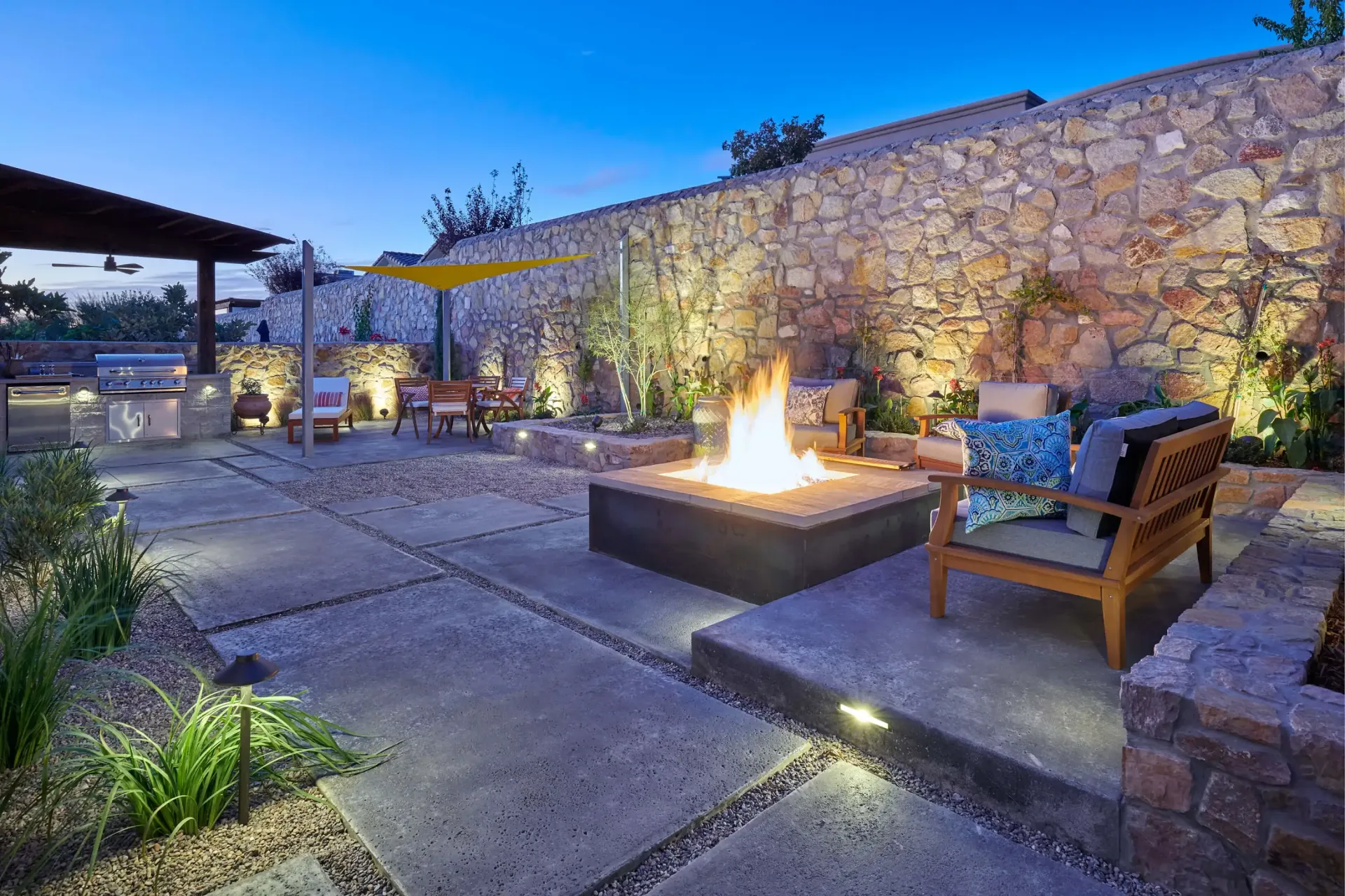 Patio with fire pit, seating, stone wall, and overhead lighting at dusk.