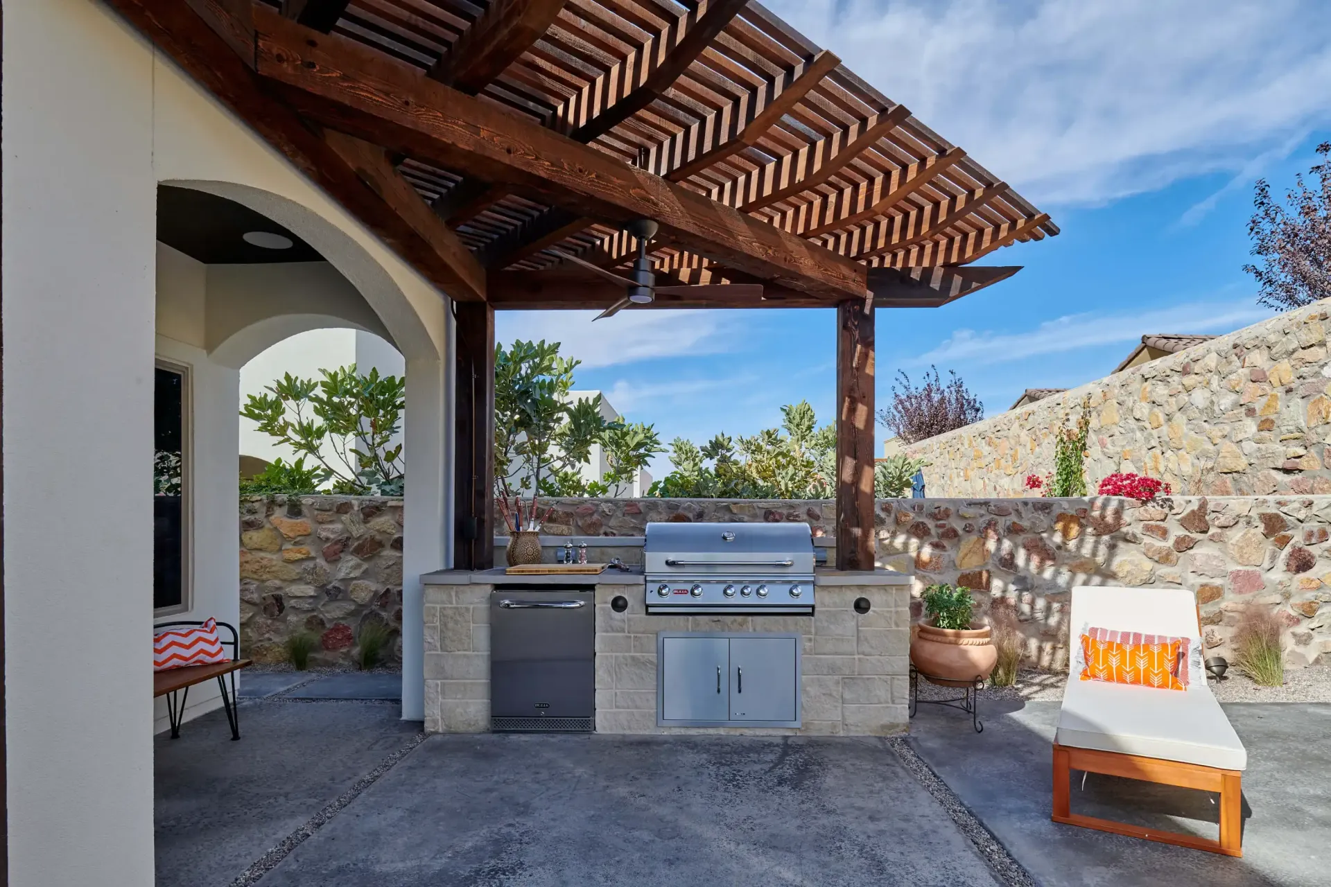 Outdoor kitchen under a wooden pergola with a grill, sink, and cabinetry. Sun lounger and stone wall visible.