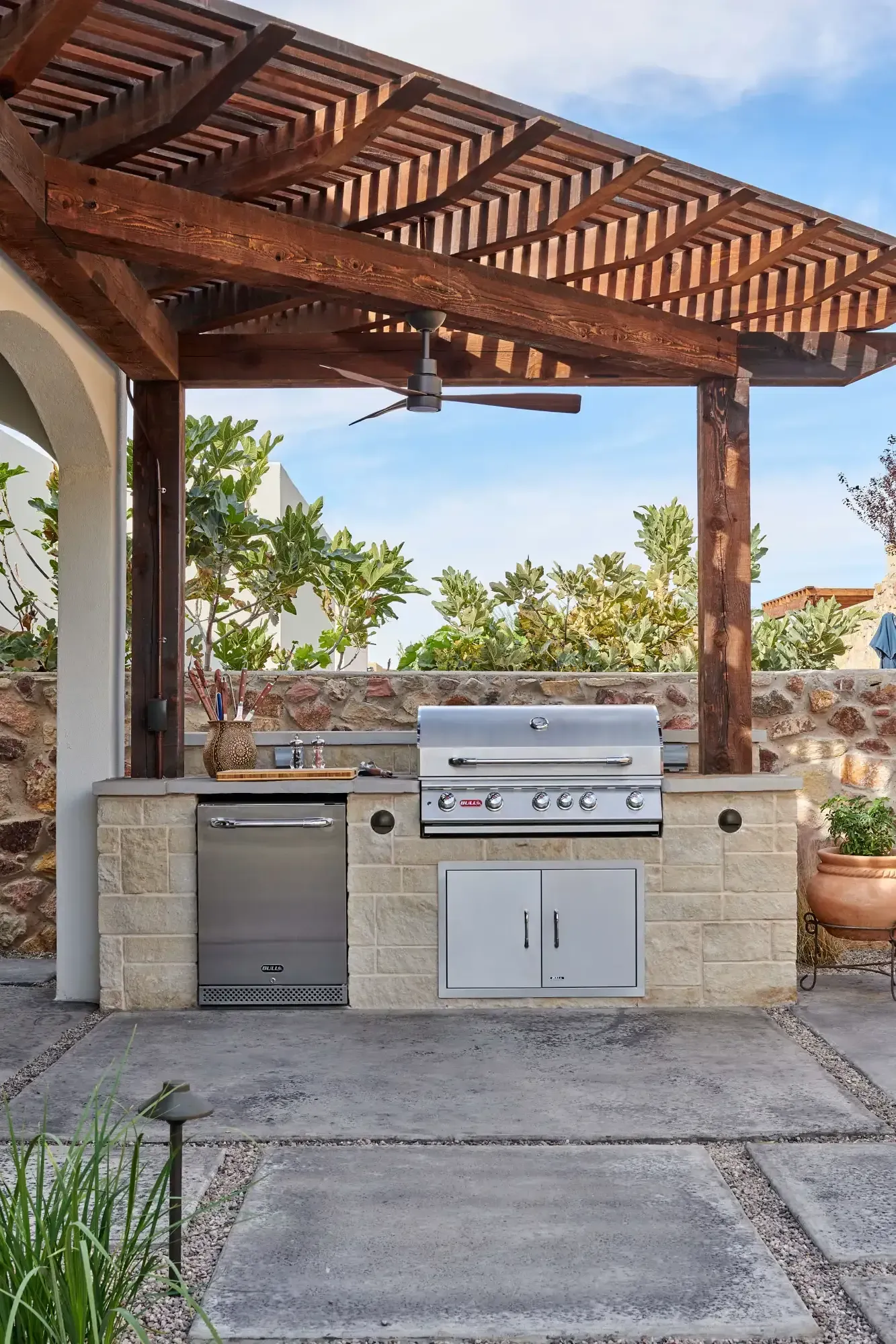 Outdoor kitchen with a grill, sink, and fridge under a wooden pergola.
