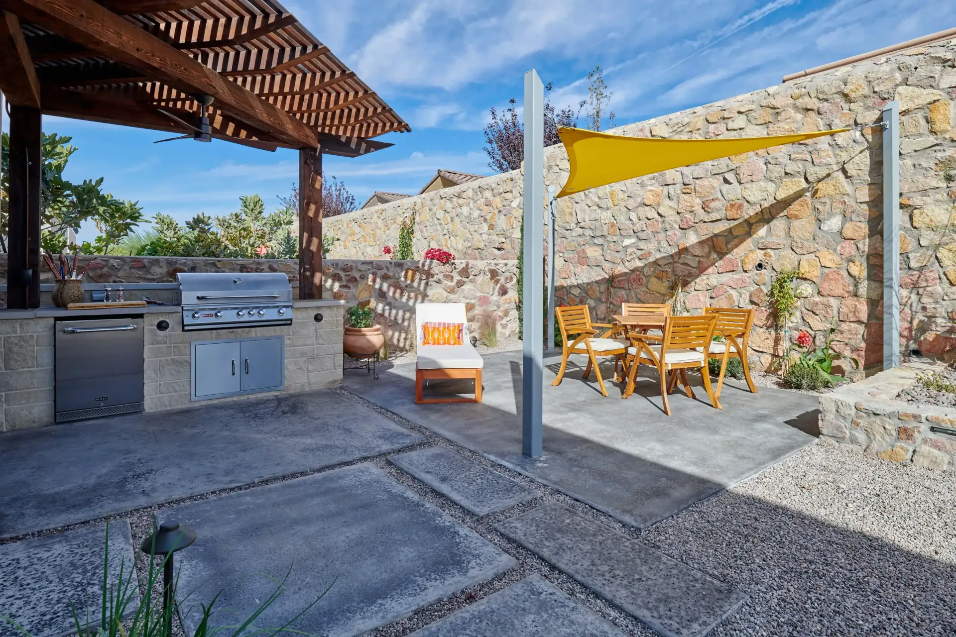 Outdoor kitchen with grill, seating, and shade structures against a stone wall. Sunny, blue sky.