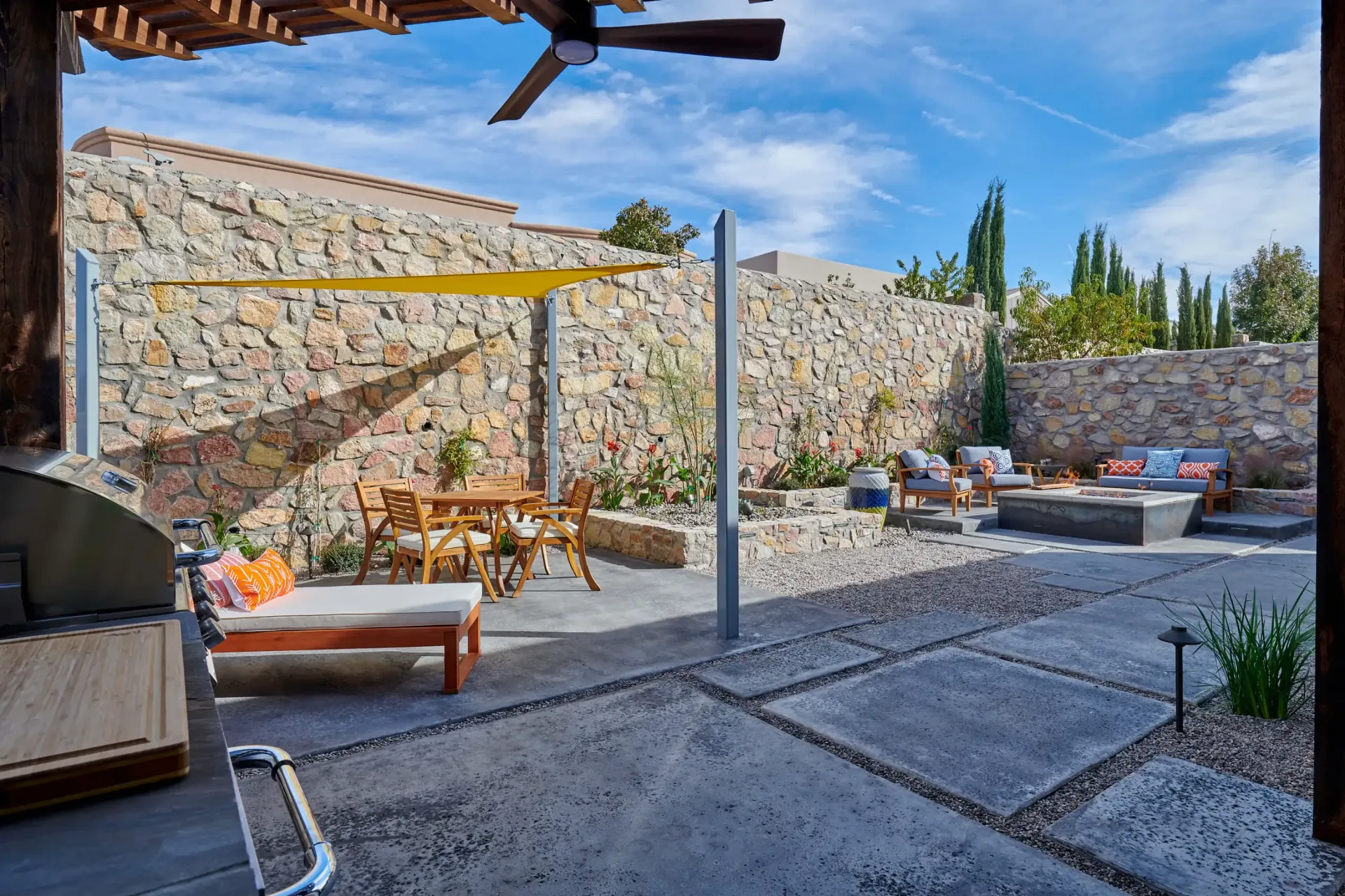 Patio with stone wall, wooden furniture, shade sail, and fire pit under blue sky.