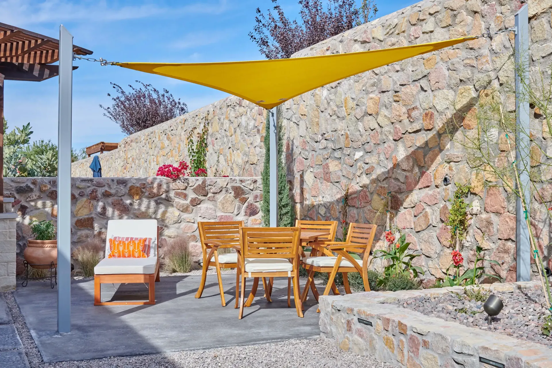 Outdoor dining area with yellow shade sail, stone wall, and wooden furniture.