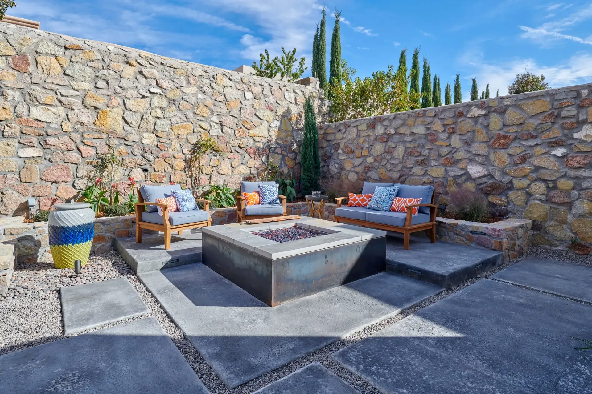 Patio with seating around a square fire pit, stone wall backdrop, blue sky.