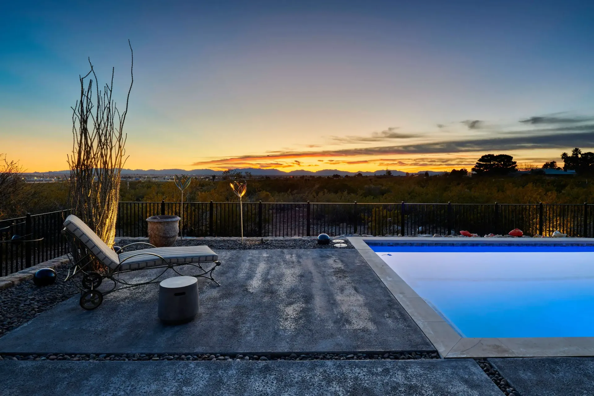 Poolside sunset view: lounge chair, small table, and pool edge overlooking a grassy landscape under a colorful sky.