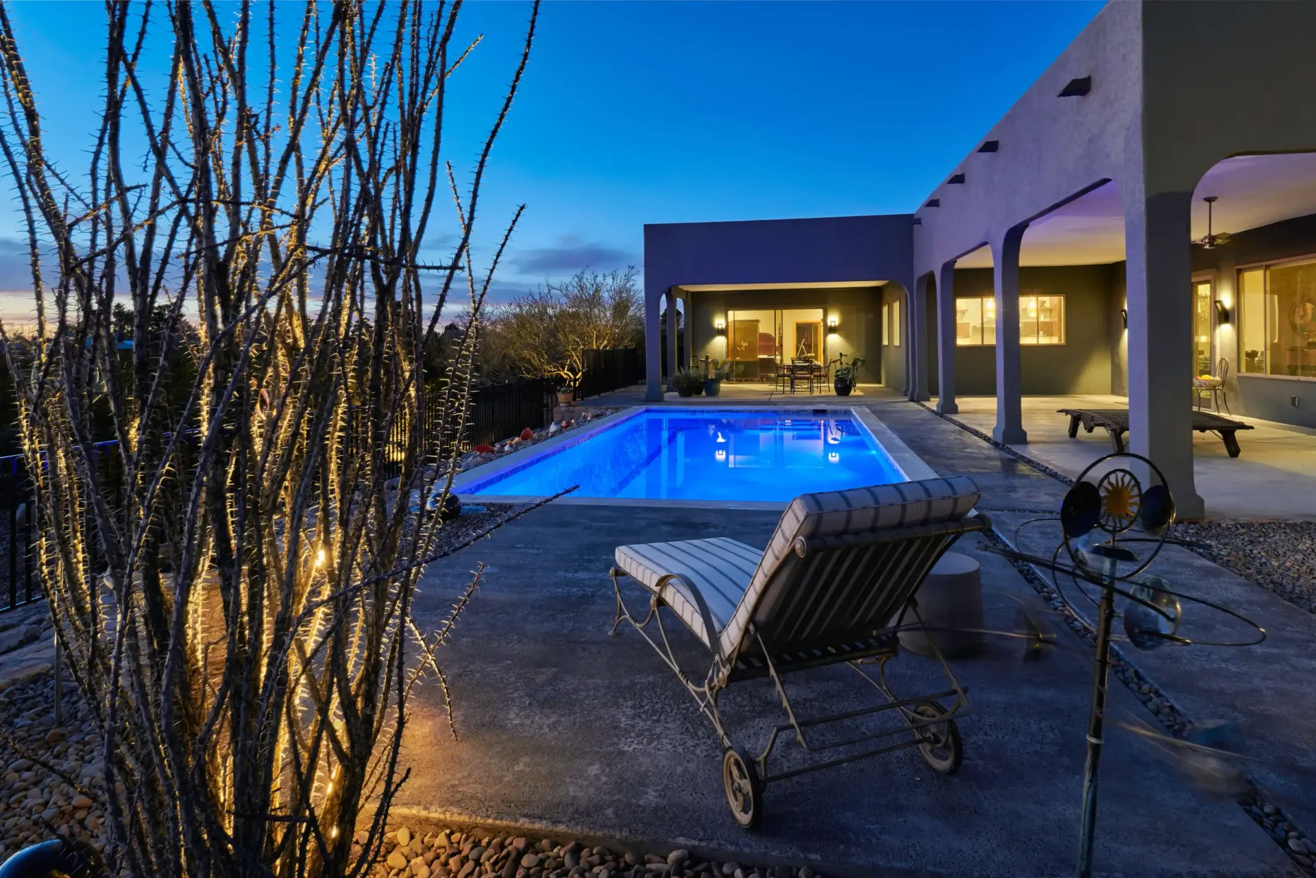 Night view of a pool and patio area, with a lounge chair and lights in front of a house at twilight.