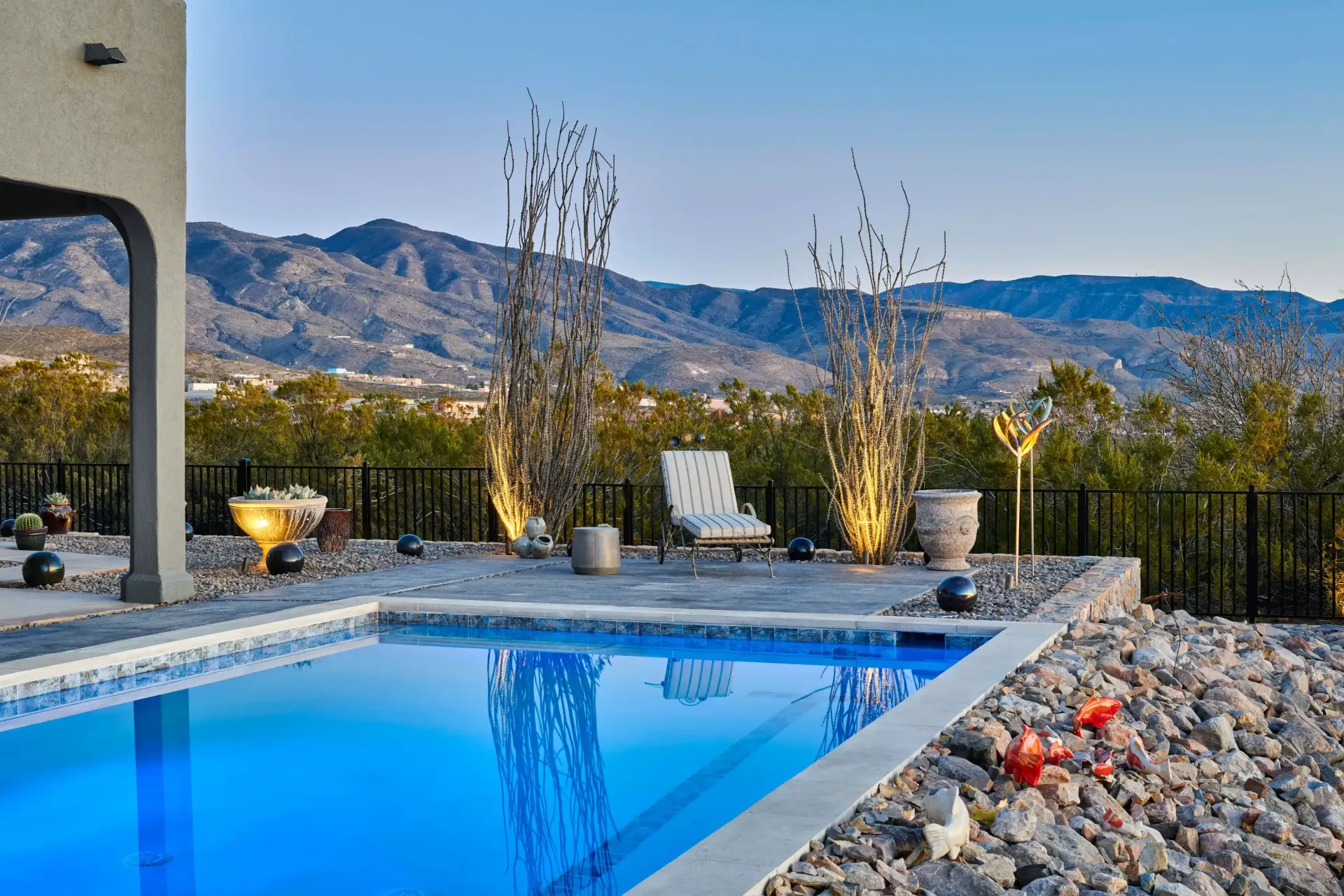 Poolside scene with pool, mountains, chair, and decorative plants. Blue water reflects sky.