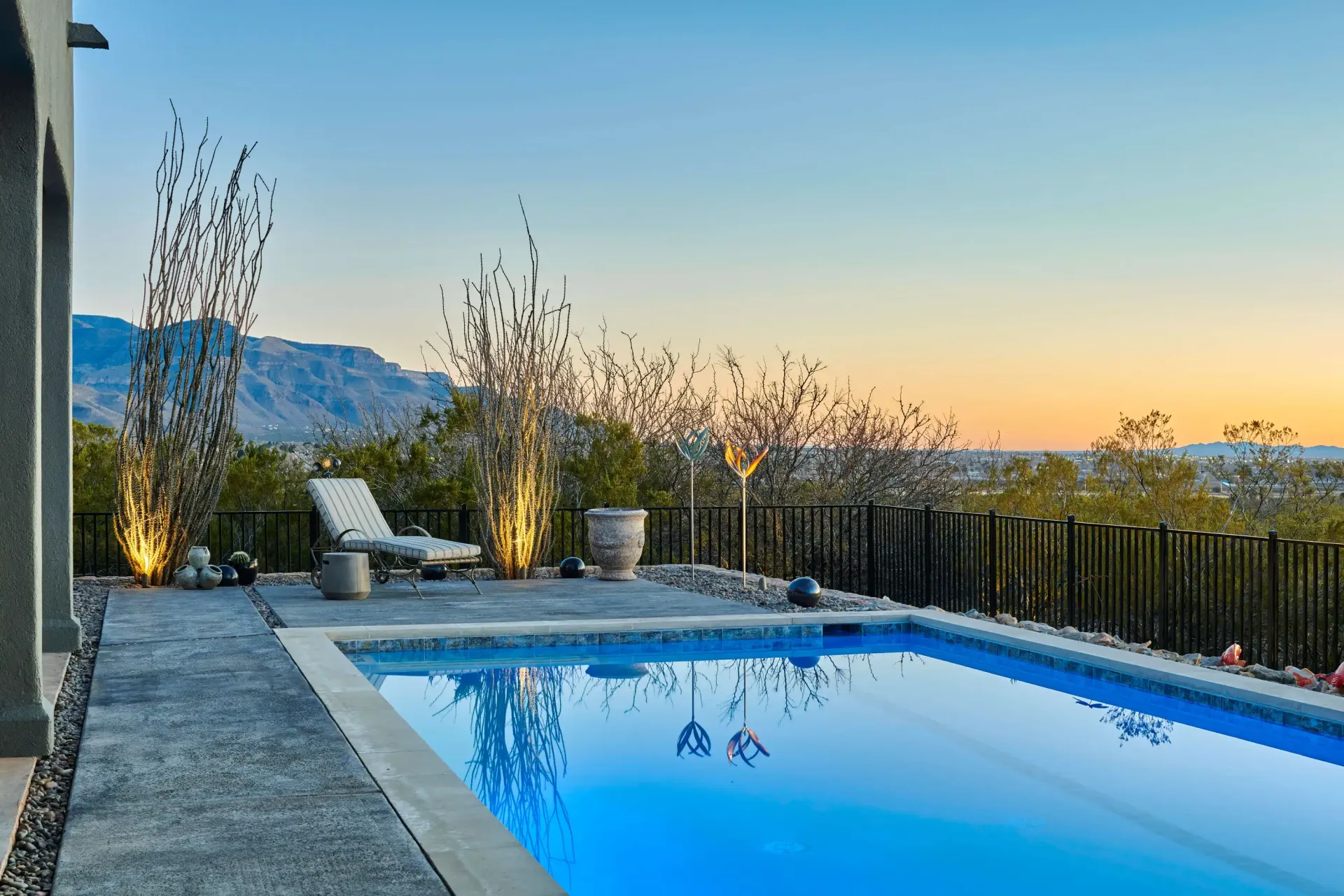 Poolside view with mountains, lounge chair, trees, and blue sky at dusk.