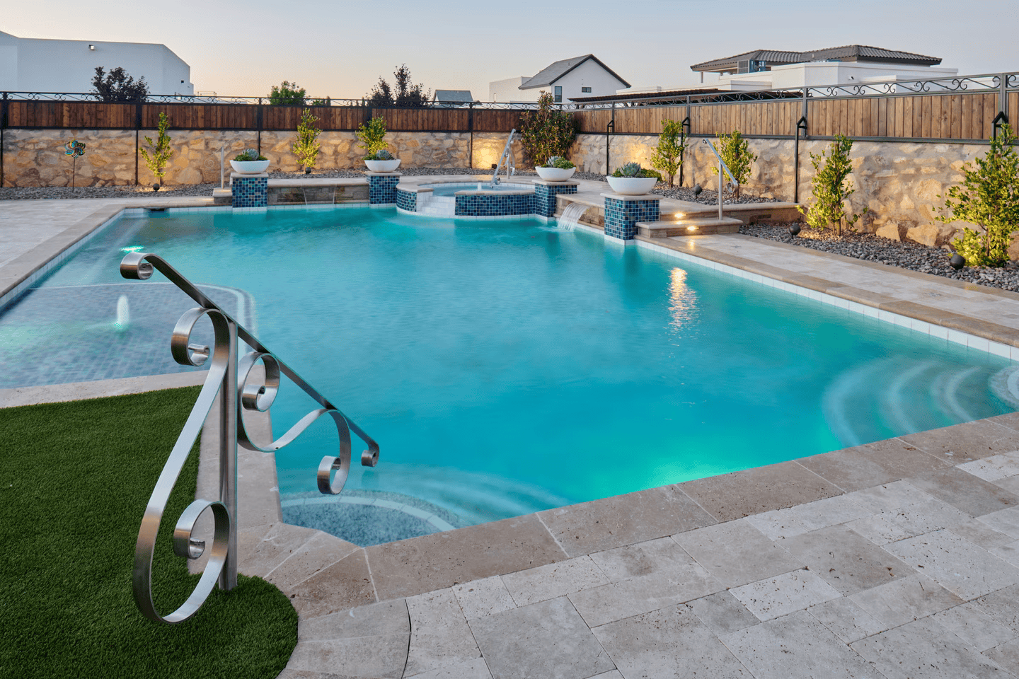 Pool with turquoise water, stone patio, green turf, and a metal handrail. Dusk lighting with lush landscaping.