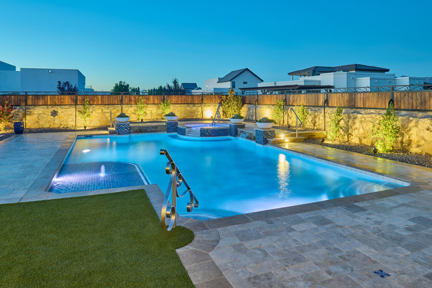 Nighttime view of a luxurious swimming pool with blue lighting, adjacent to a spa, grass, and stone patio.
