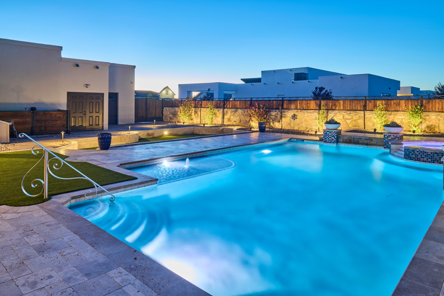 Pool lit with blue lights, patio with stone tiles, and a white stucco building in the background.