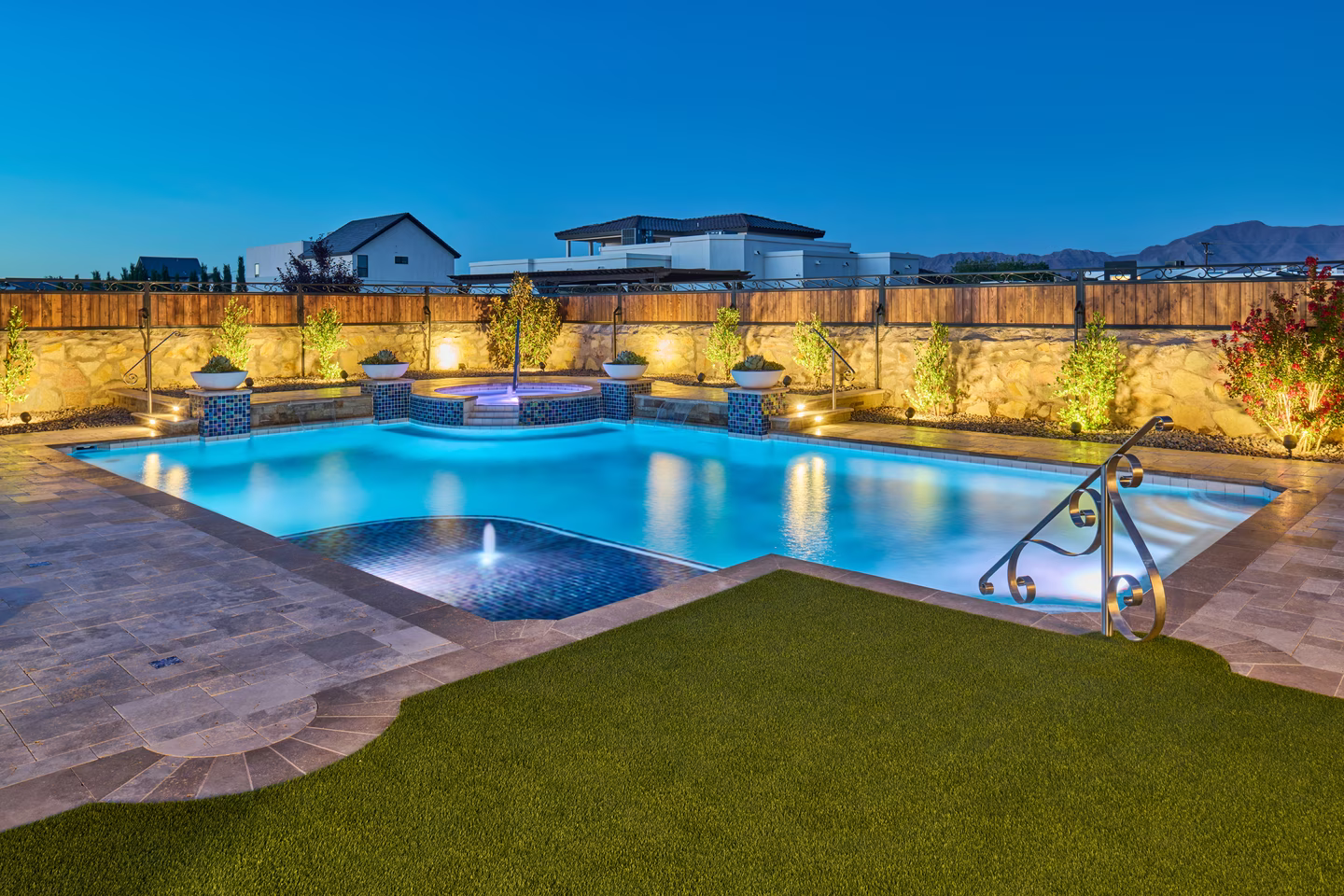 Pool area at dusk with illuminated pool, landscaping, and house in the background.
