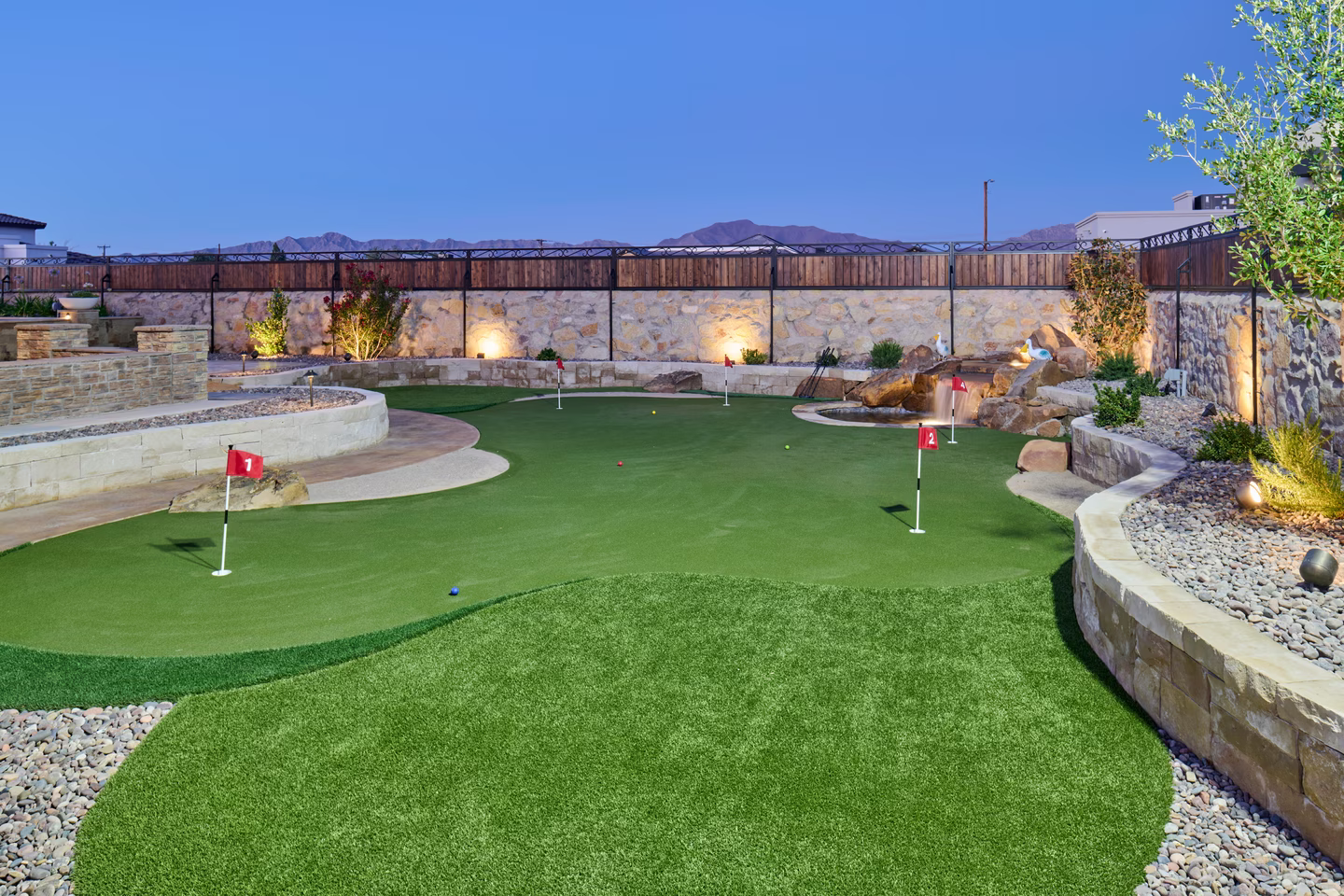 A backyard putting green with red flags, surrounded by stone walls and landscaping at dusk.