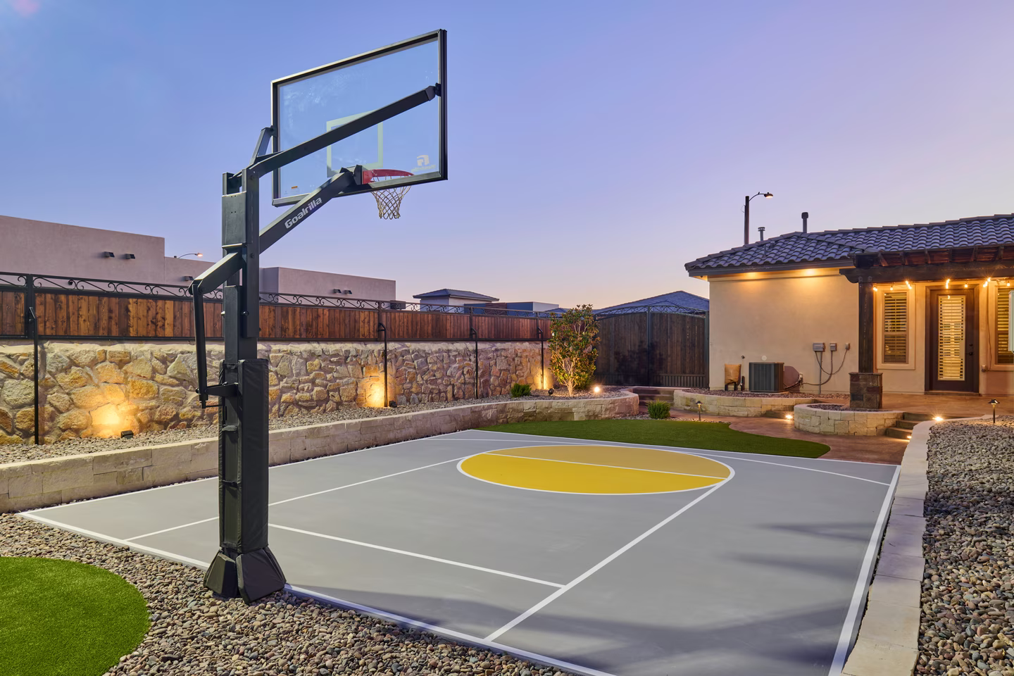 Basketball court in backyard with hoop, gray court, yellow circle, green grass, and house.