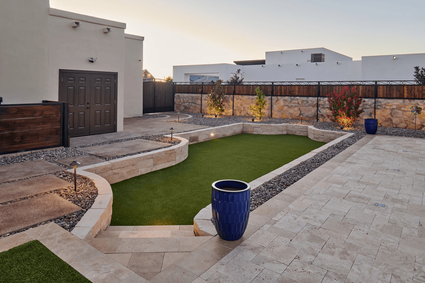 A well-landscaped backyard featuring a raised artificial lawn, stone walls, and blue pottery against a white building.