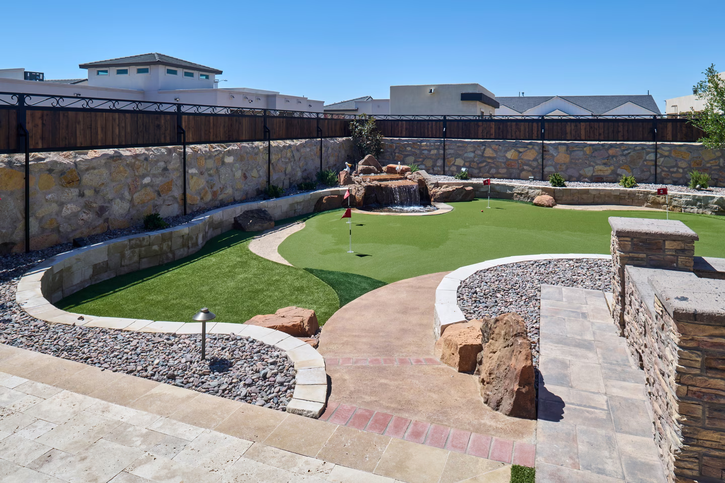 Backyard putting green with waterfall feature, stone walls, and flagsticks.