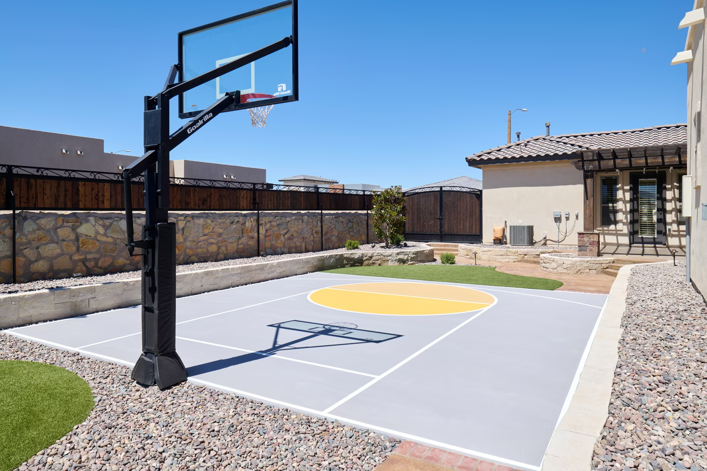 Basketball court in backyard, gray surface with yellow circle. Black hoop and post. Sunny, blue sky.
