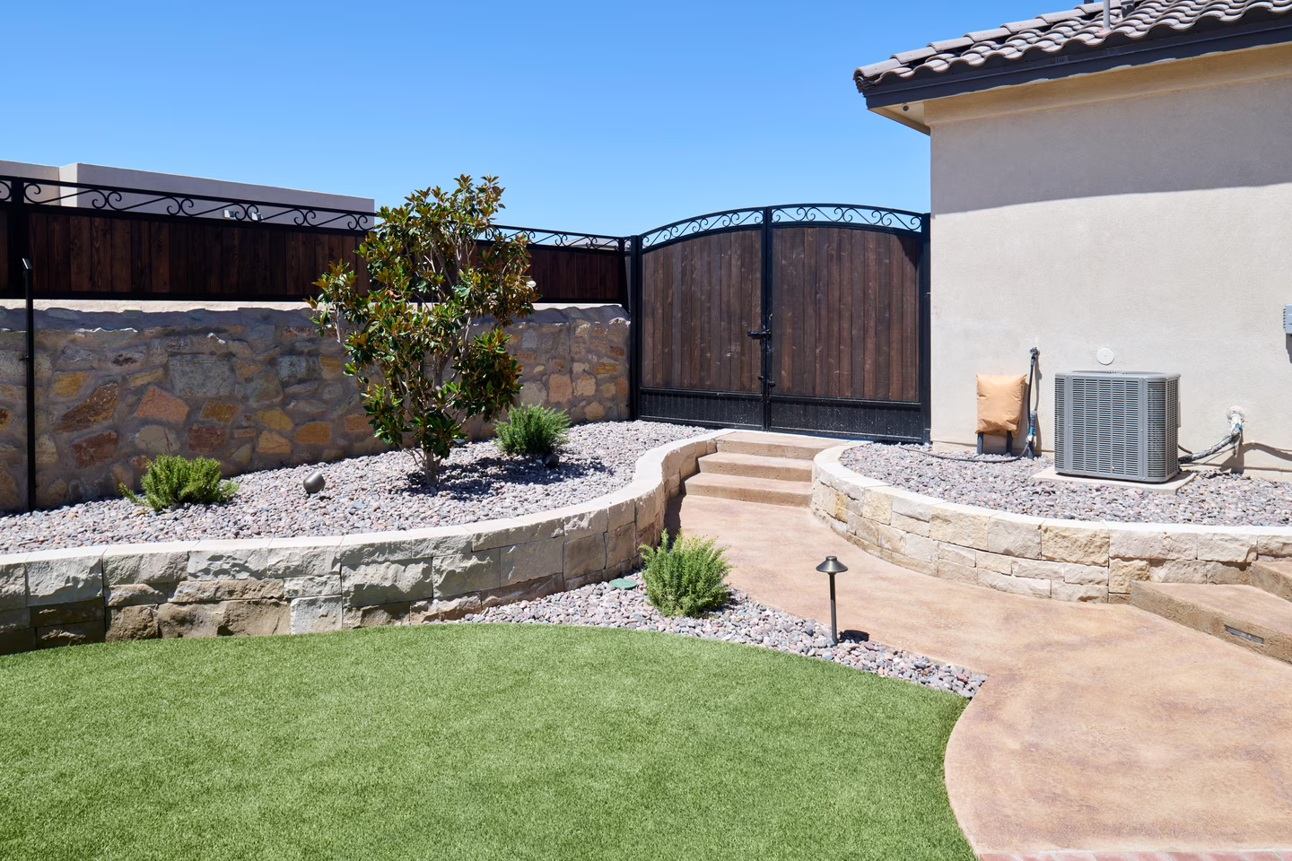 Backyard with stone walls, stairs, and path. Features green grass, gravel, and a tree.