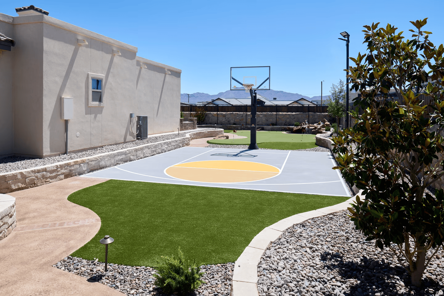 Outdoor basketball court with gray and yellow painted surface, surrounded by green turf and rock landscaping.