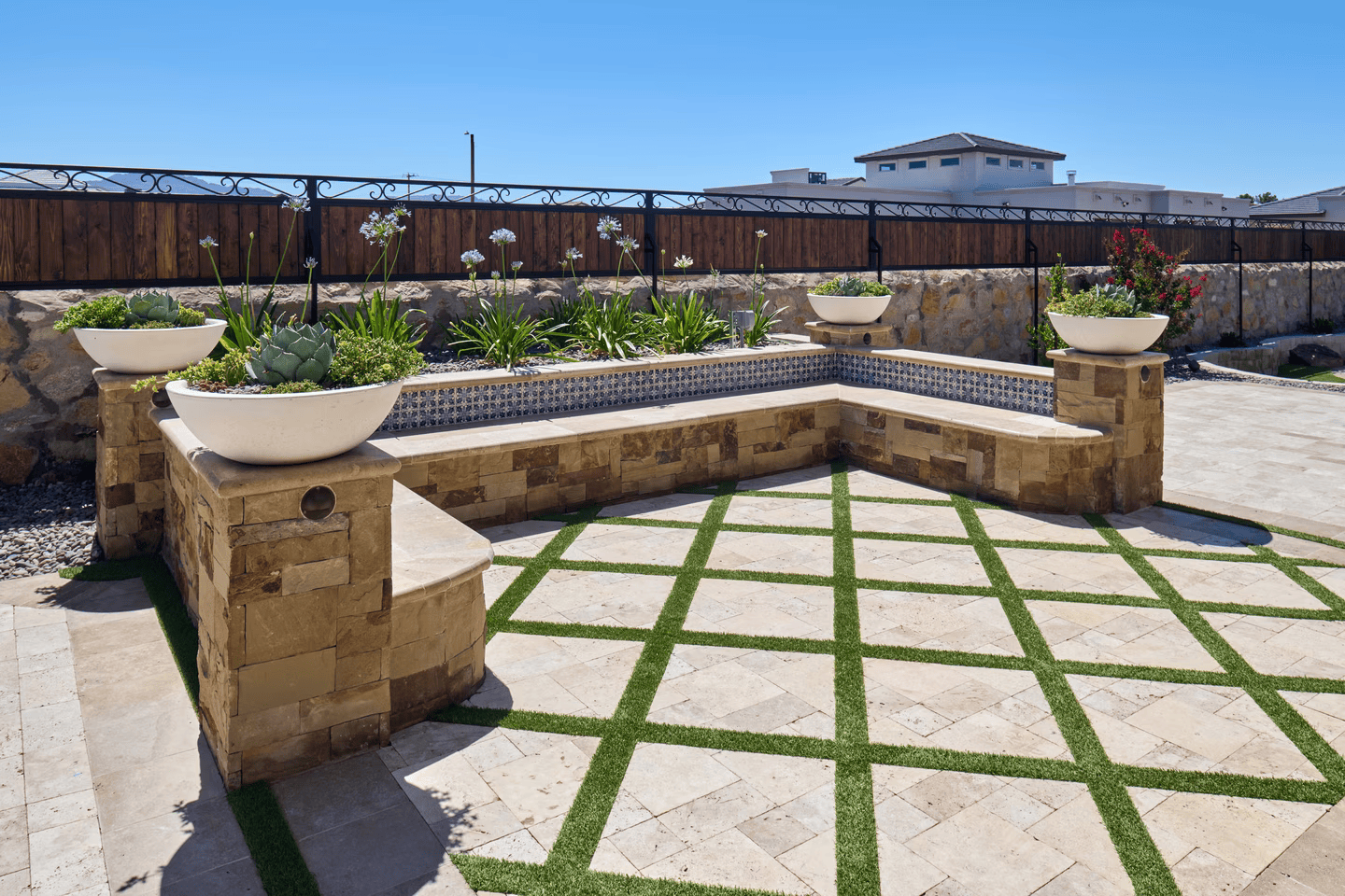 Stone bench with planters in a landscaped yard. Green grass squares and blue sky.