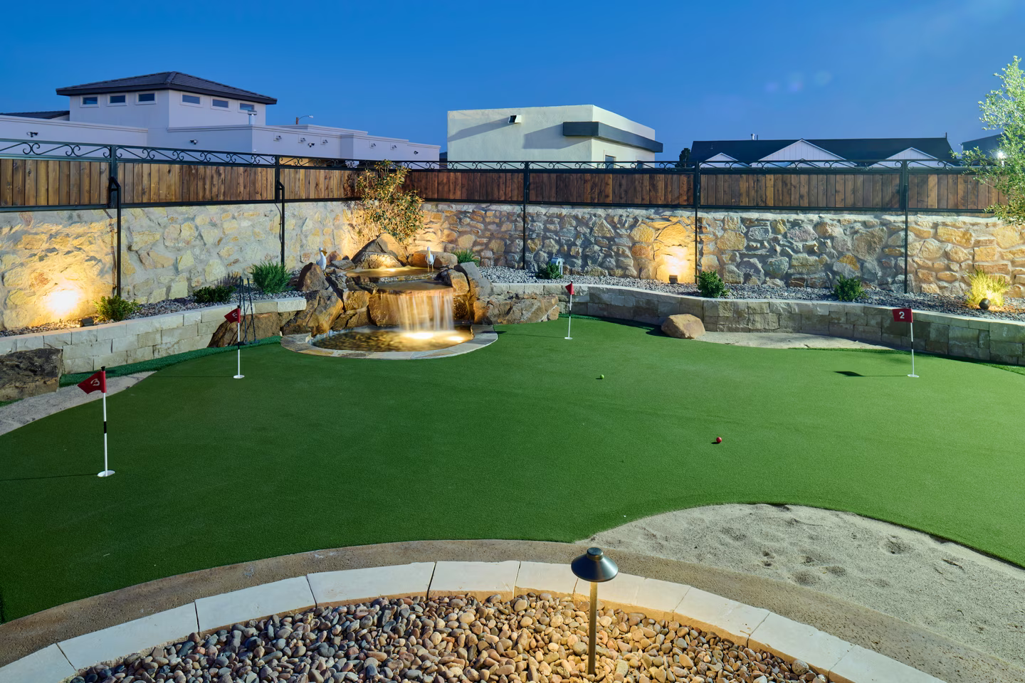 Miniature golf course with waterfall feature, illuminated at dusk, surrounded by stone and wooden fence.