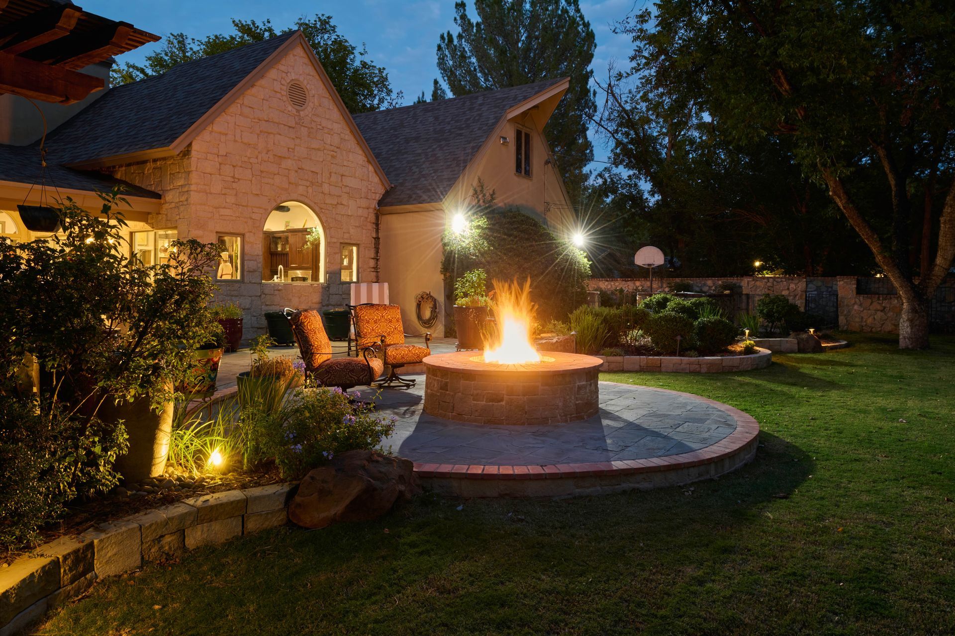 Backyard with a lit fire pit and patio seating at dusk. House and landscaping are lit by warm lights.