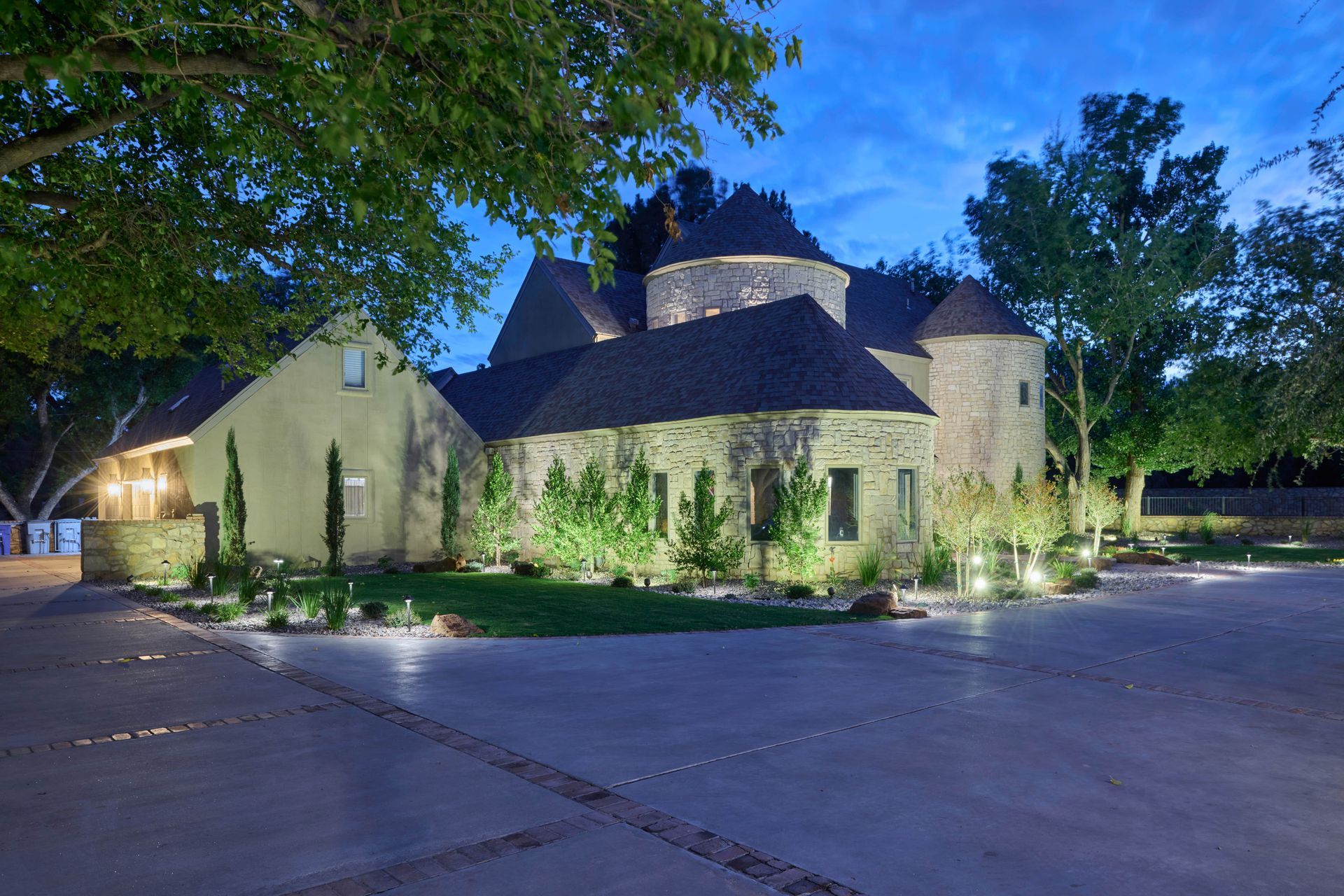 Stone castle-like building illuminated at dusk with trees and a paved driveway.
