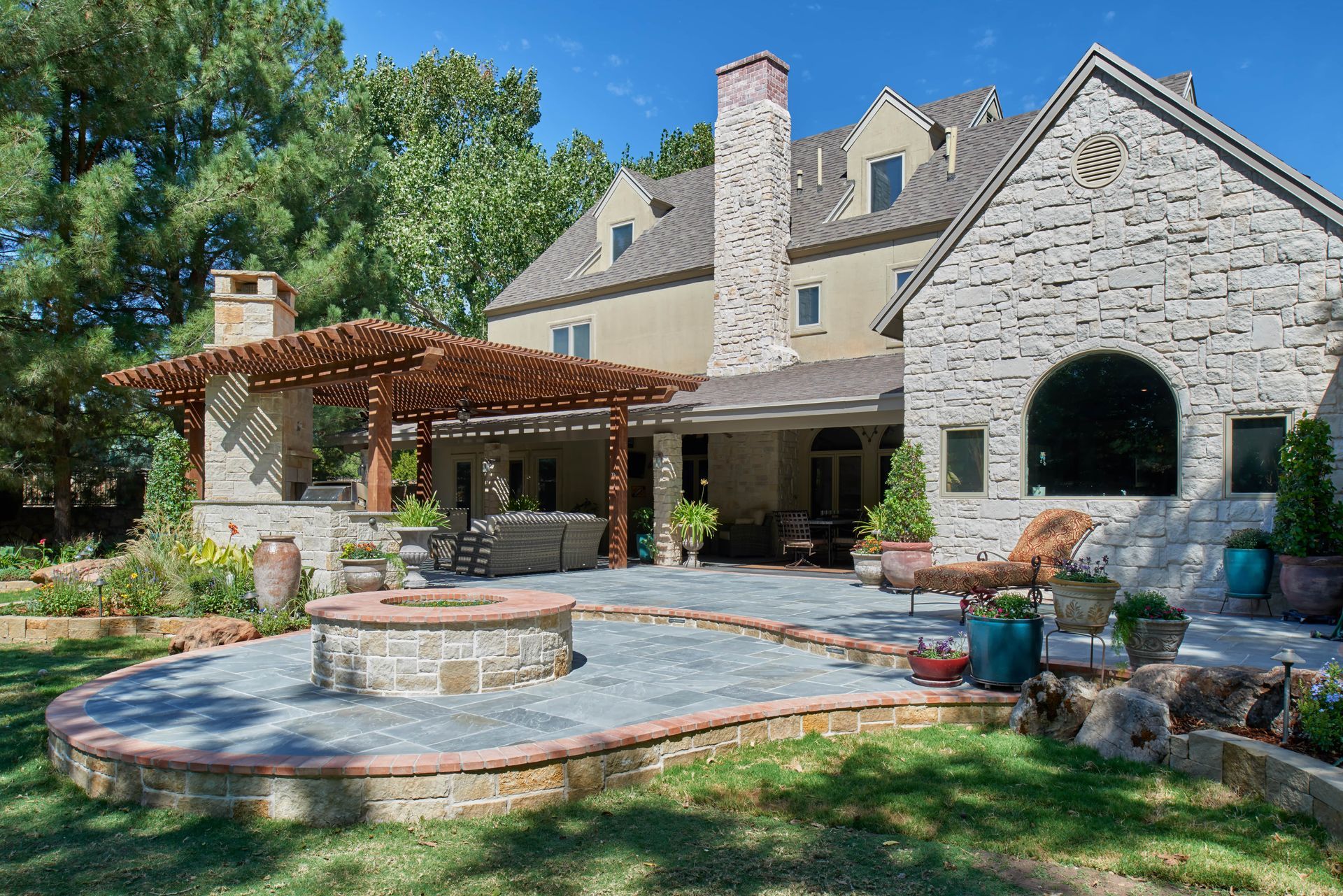 Backyard patio with fire pit, pergola, stone walls, and house. Blue sky and green yard.