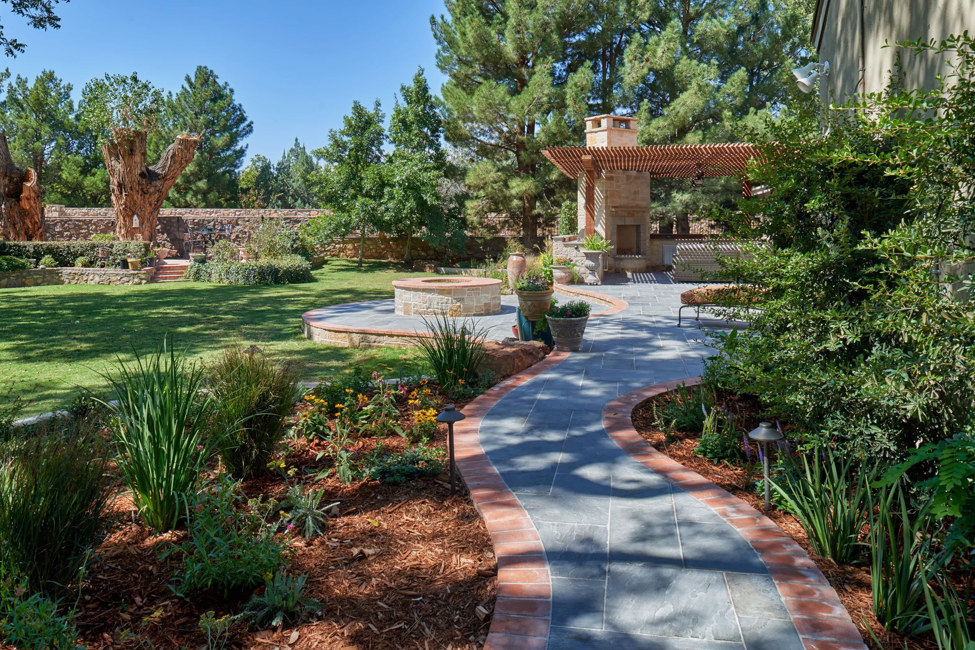 Stone patio and path in a landscaped backyard with a fire pit, and a pergola-covered outdoor kitchen.