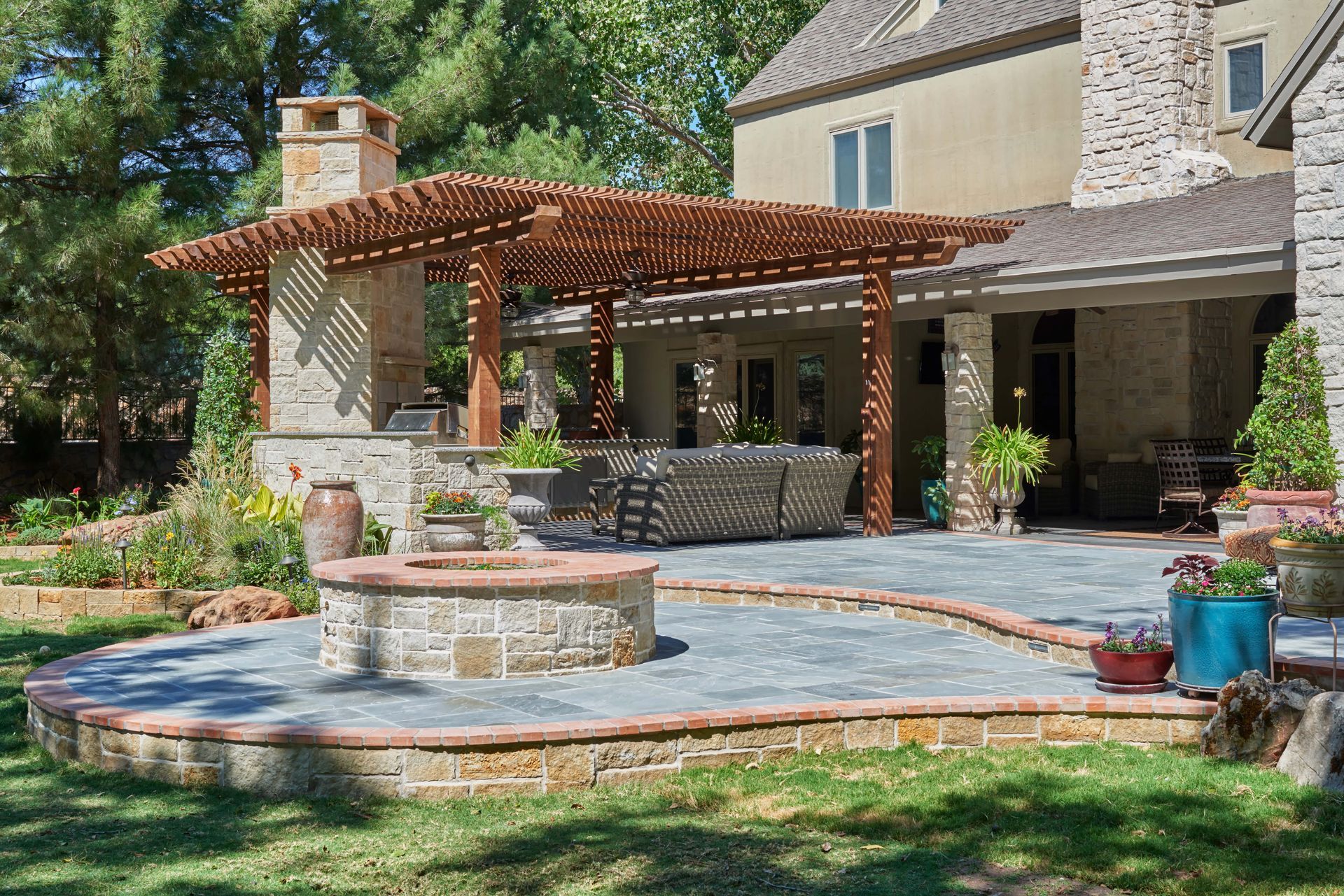 Backyard patio with fire pit, pergola, stone features, and seating. Lush green landscaping and a sunny day.