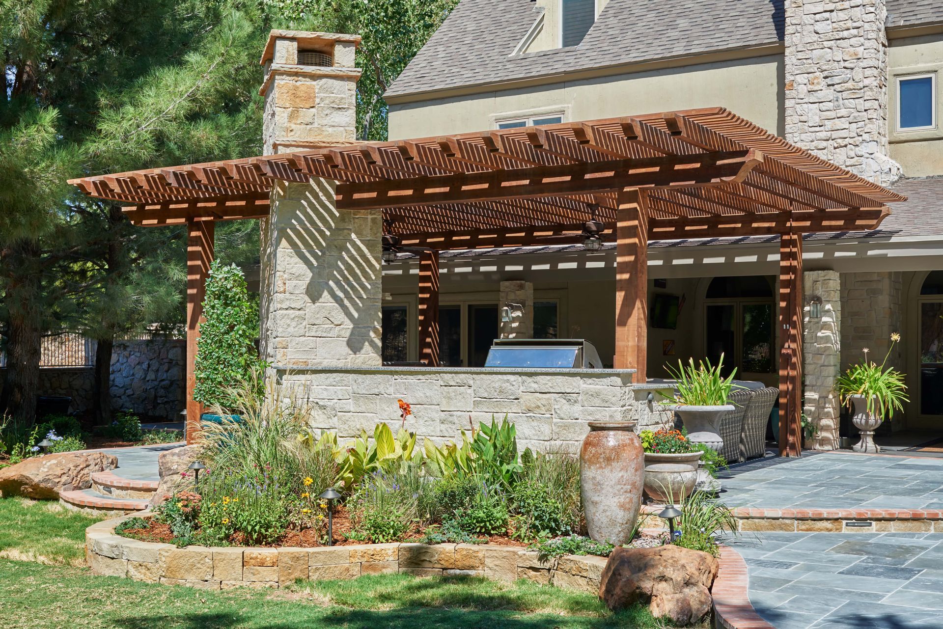 Pergola with stone pillars and a built-in grill, attached to a house with landscaping.