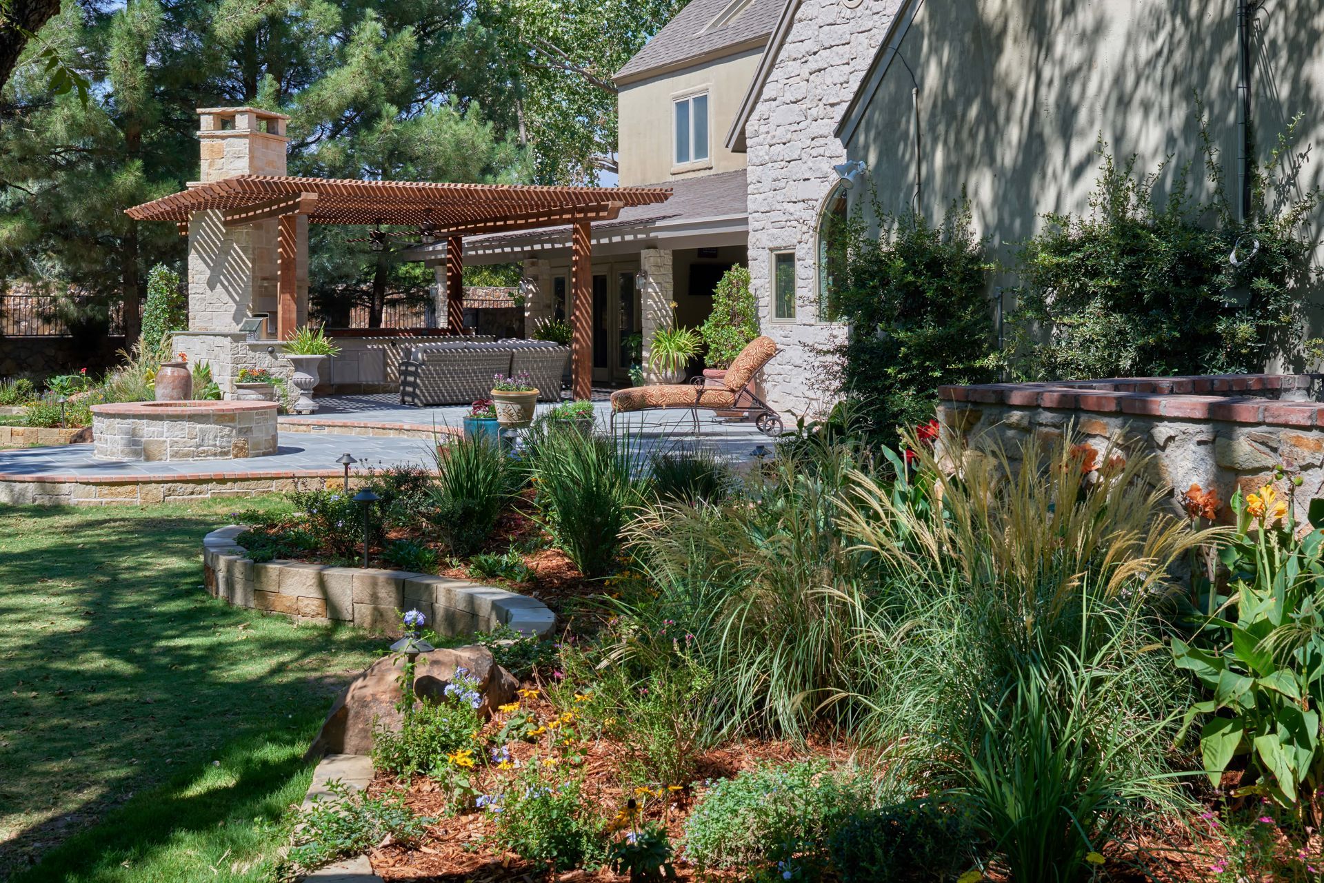Stone patio and pergola with fireplace, lush landscaping, and a house in the background.