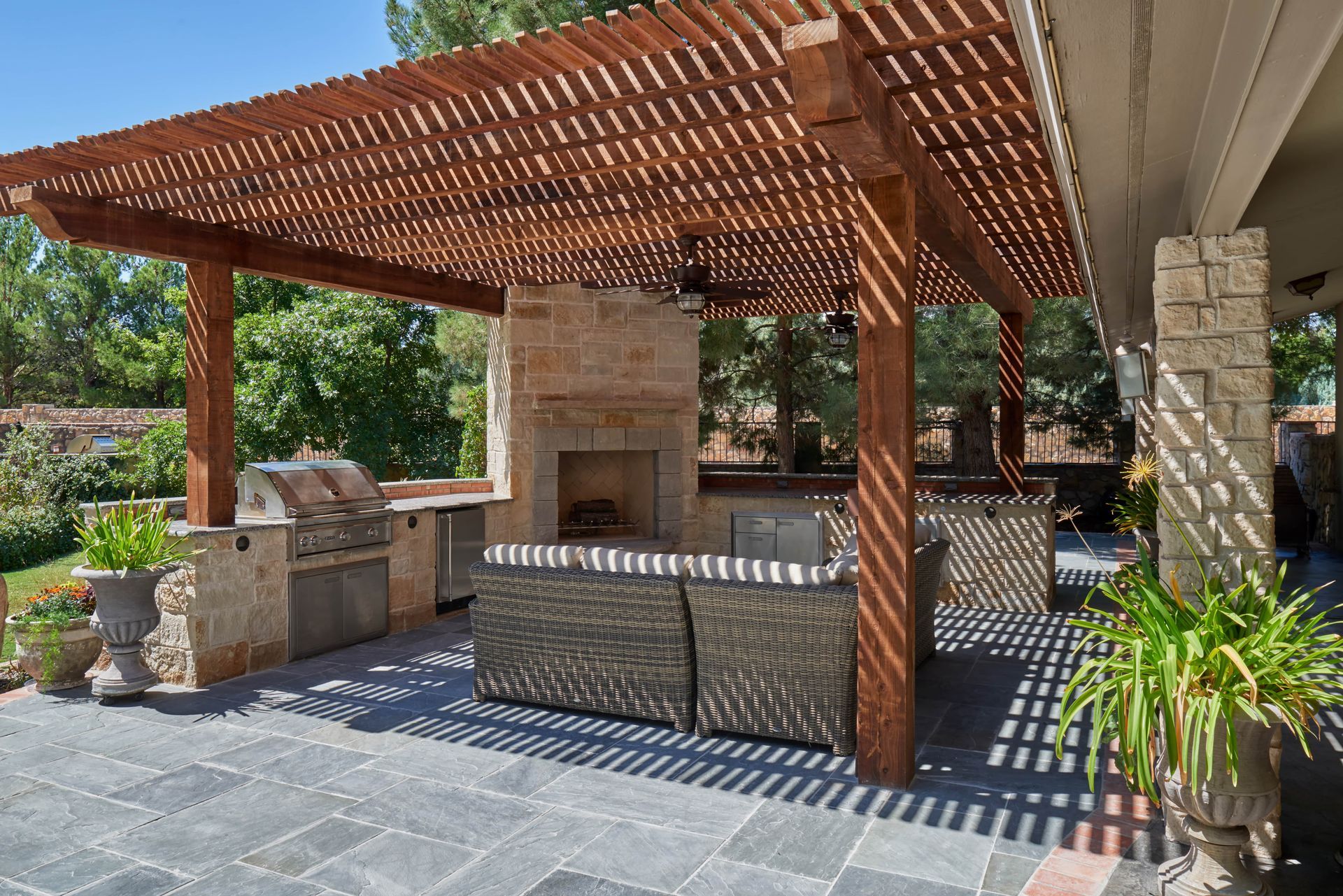 Outdoor patio with pergola, fireplace, grill, and seating. Sunlight casts shadows on the stone and dark pavers.