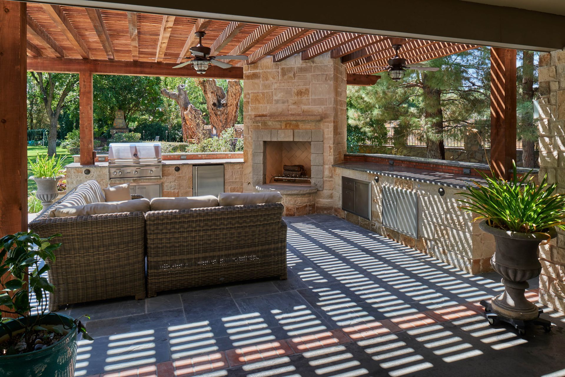 Outdoor patio with stone fireplace, grill, seating, and overhead wooden trellis.