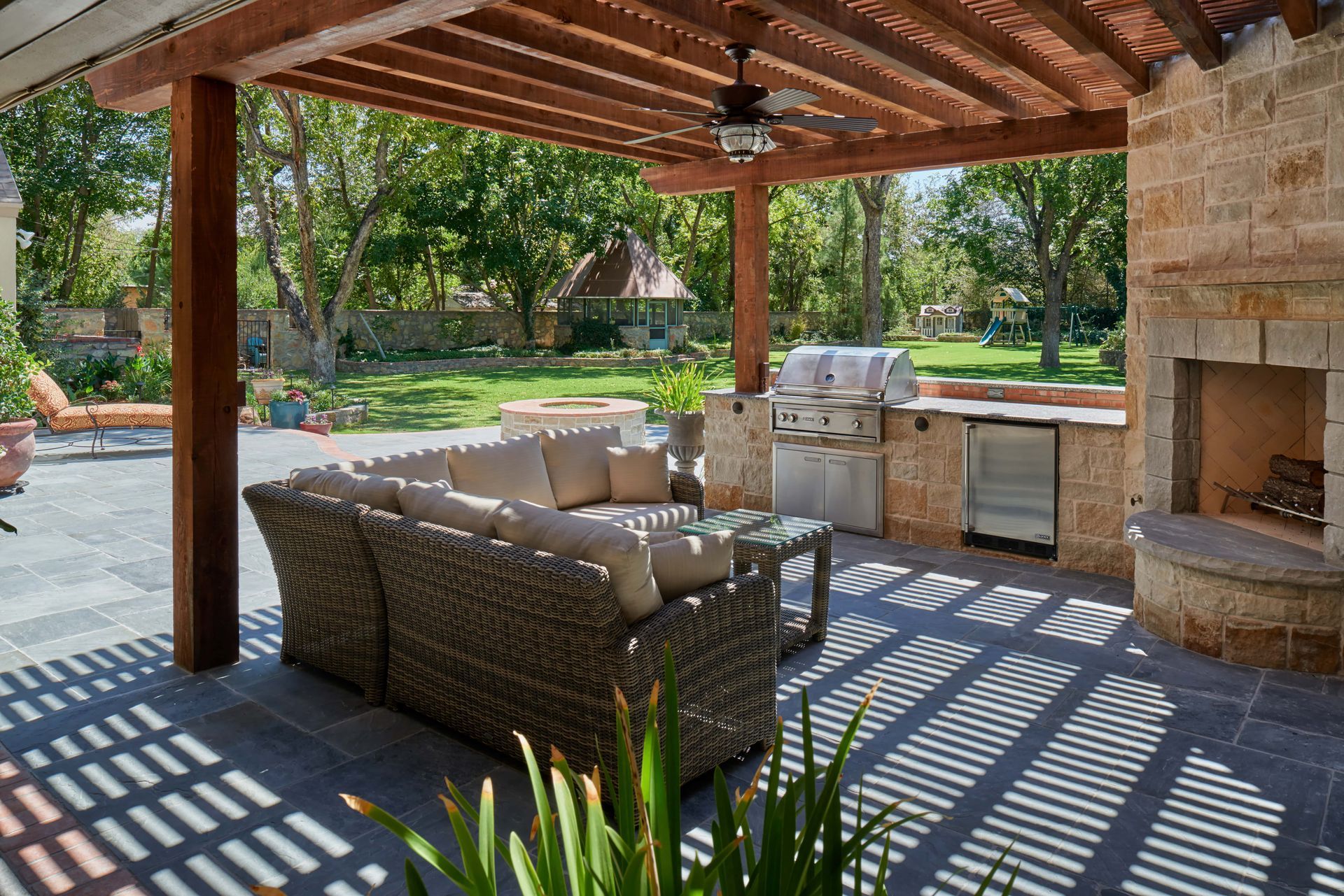 Outdoor patio with sectional sofa, grill, fireplace, and pergola, in a backyard setting.