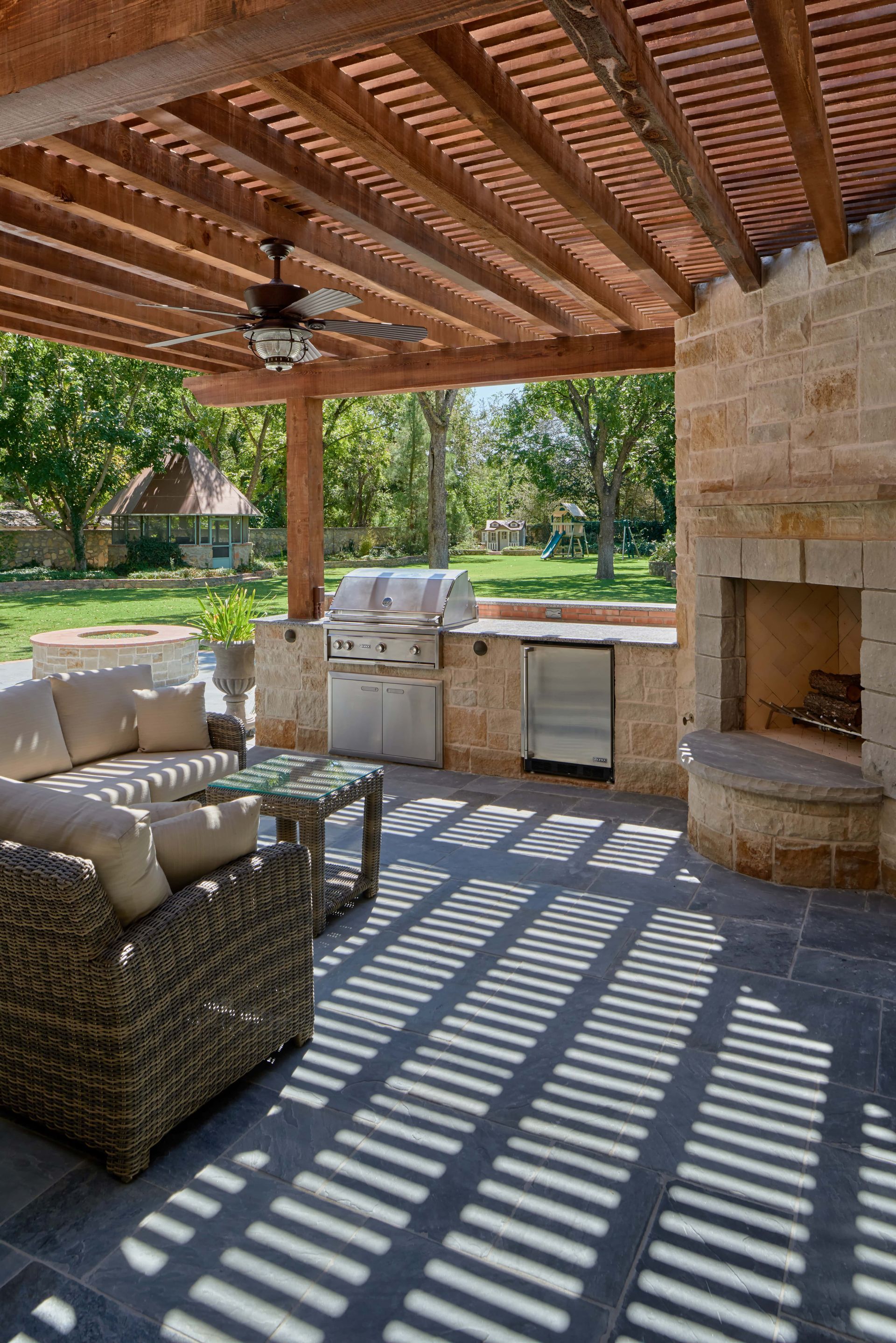 Patio with a pergola, outdoor kitchen, fireplace, and seating area. Sunlight creates striped shadows.