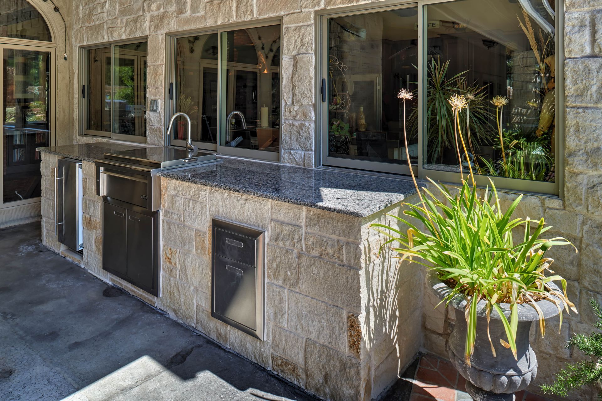 Outdoor kitchen with a sink, grill, and storage built into a stone structure next to windows.