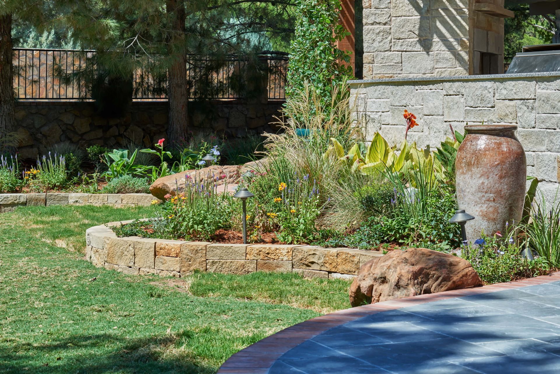 Stone-walled garden bed with varied plants, a large pot, and grass. Sunny outdoor setting.