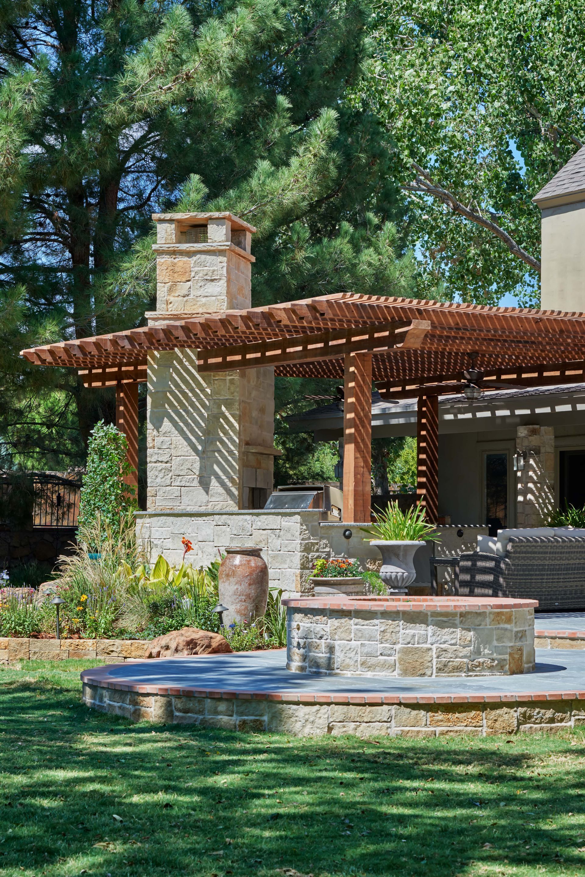 Stone patio with outdoor fireplace, pergola, and tiered fountain. Lush green lawn and trees in the background.