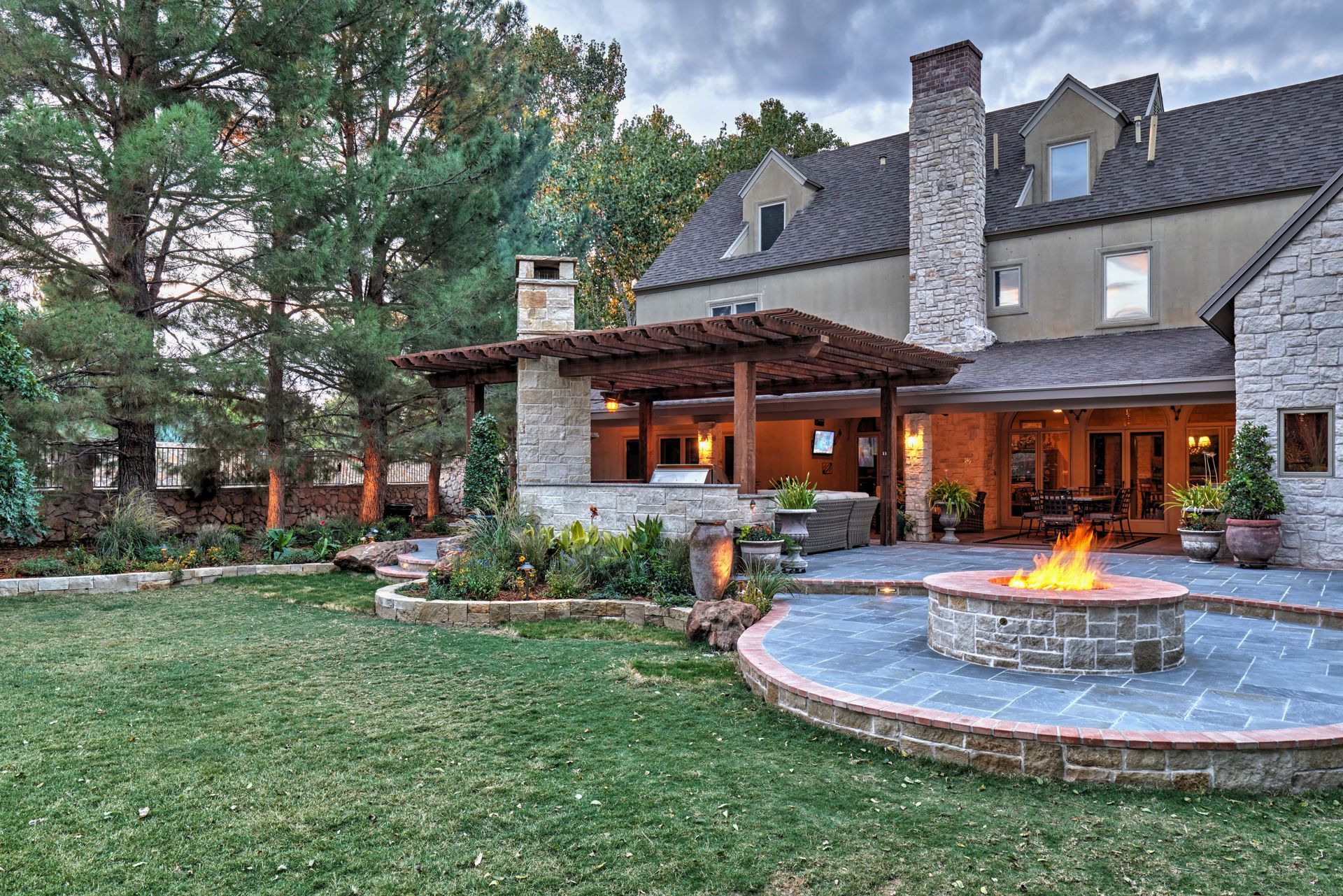 Backyard with a stone patio, fire pit, pergola, and large house. Overcast sky.