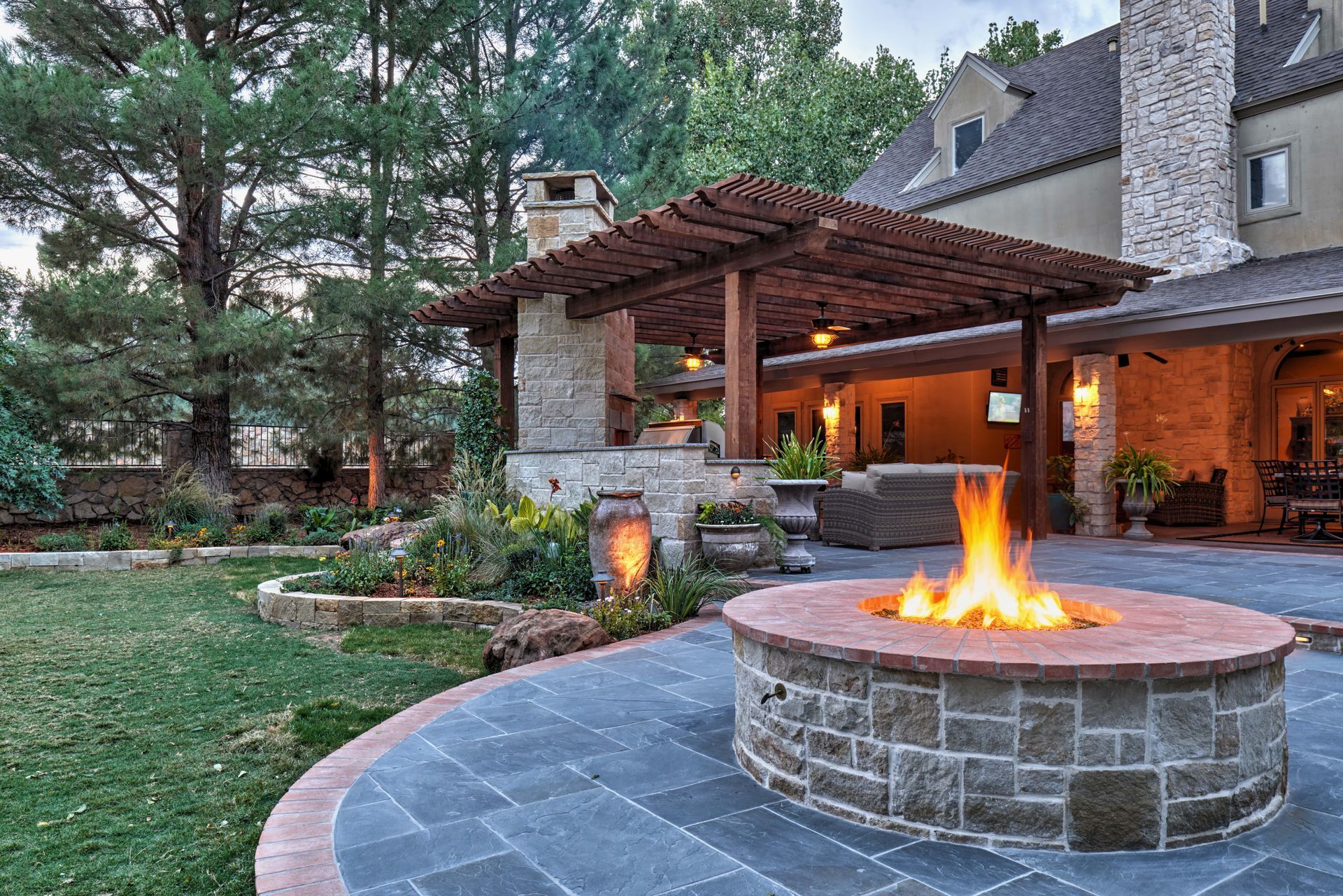 Stone patio with fire pit, pergola, and house. Fire burns in pit, evening setting.