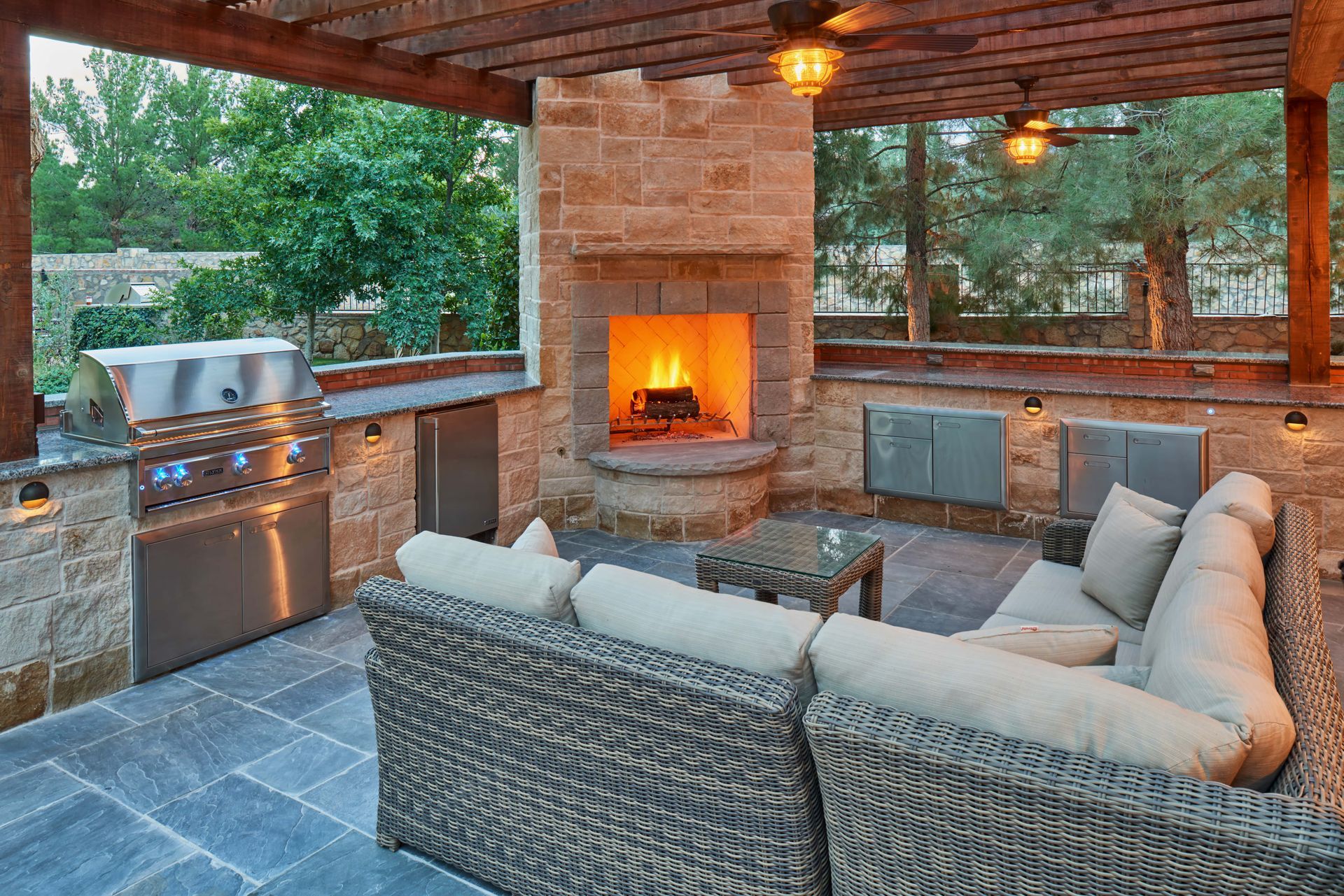 Outdoor kitchen with a stone fireplace, grill, seating, and wooden pergola.