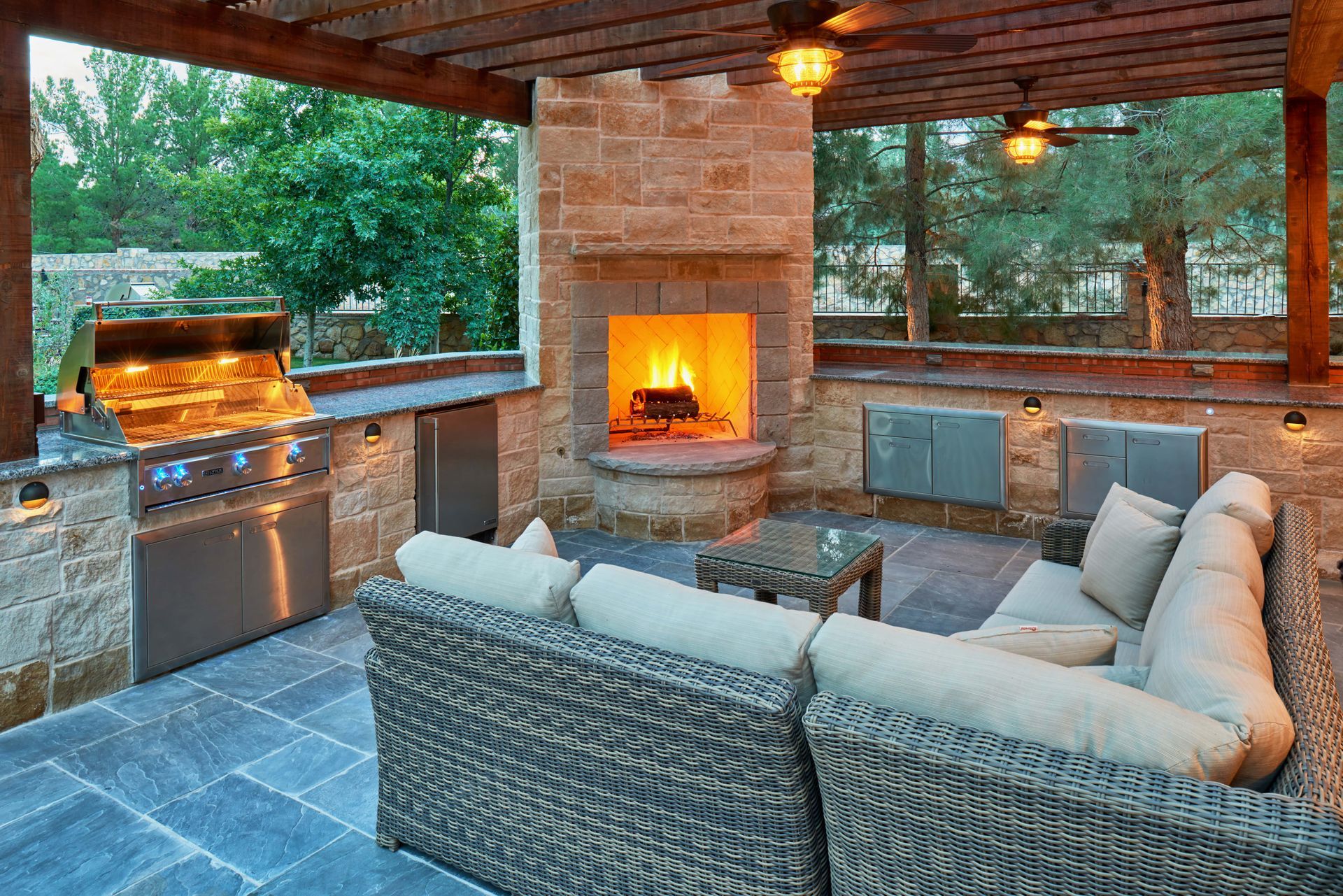 Outdoor kitchen with stone walls, fireplace, grill, and wicker seating under a wooden pergola.