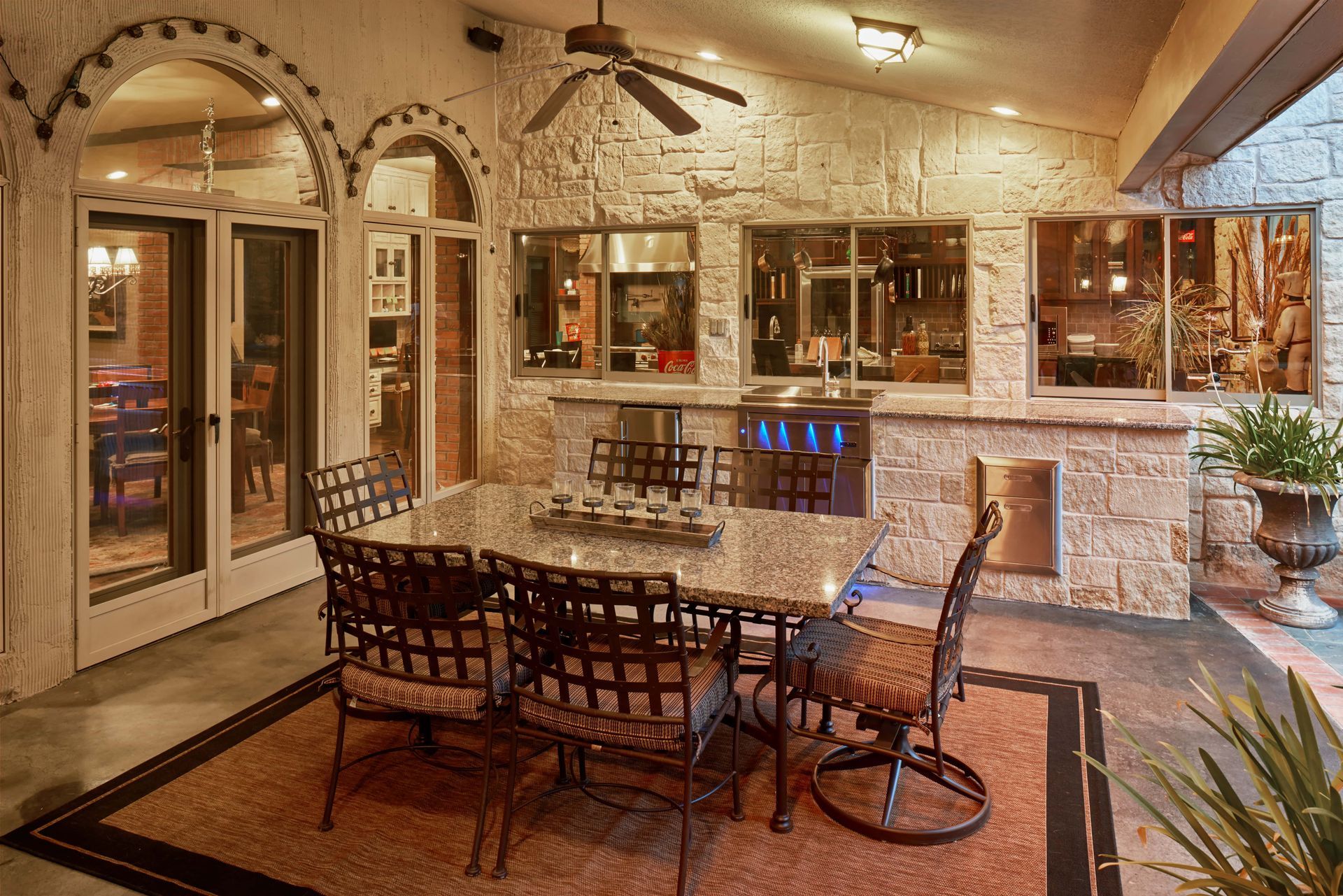 Patio with stone wall, dining table, and outdoor kitchen. Beige stone, dark table and chairs, rug, and windows.