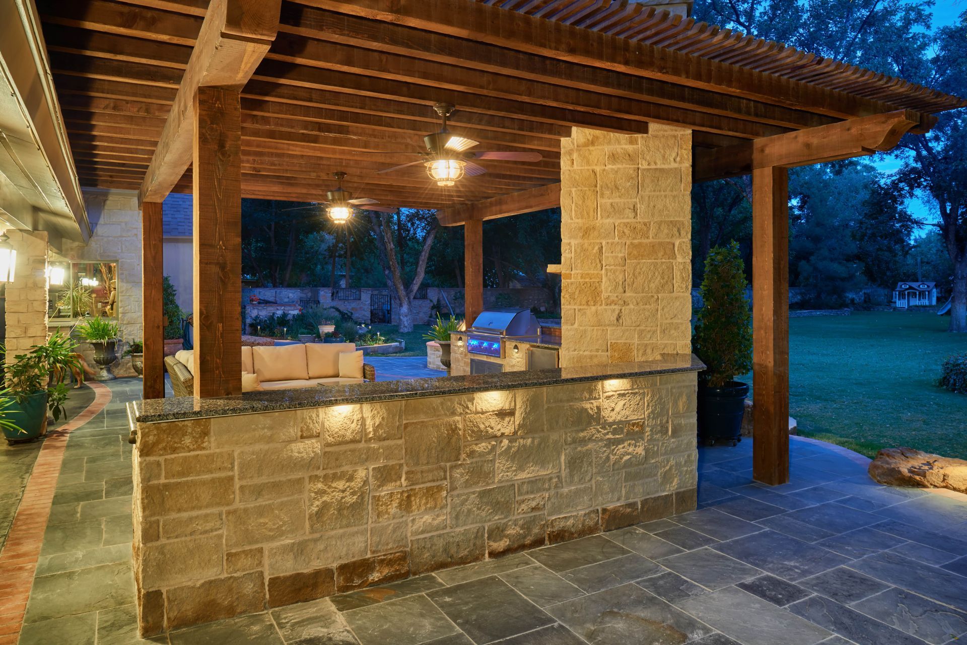 Outdoor kitchen with stone bar, wooden pergola, and blue slate patio at dusk.